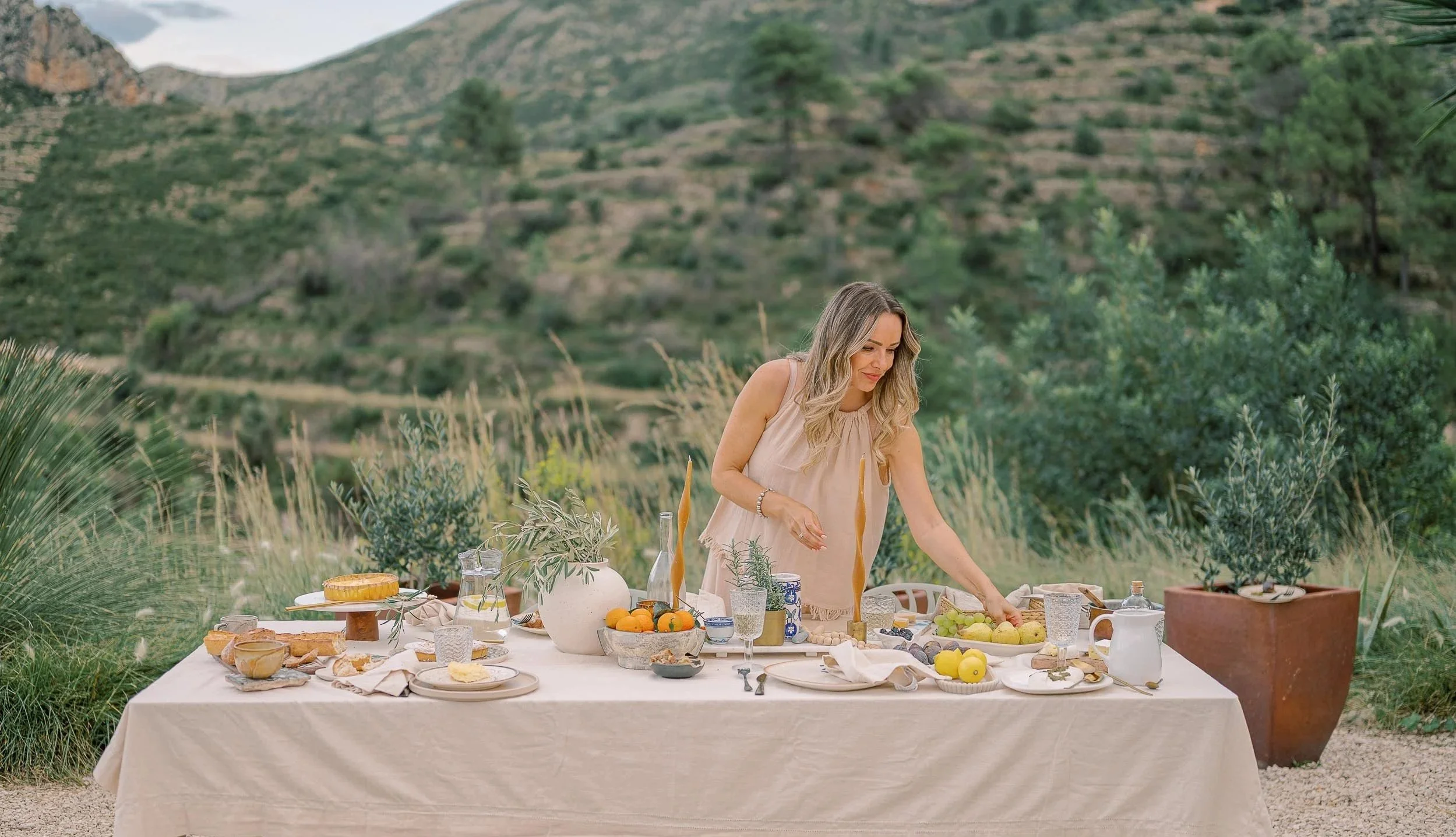 Denisa setting a table outdoors in a mountainous landscape with greenery and desert plants, decorated with candles, vases, fruits, and tableware.