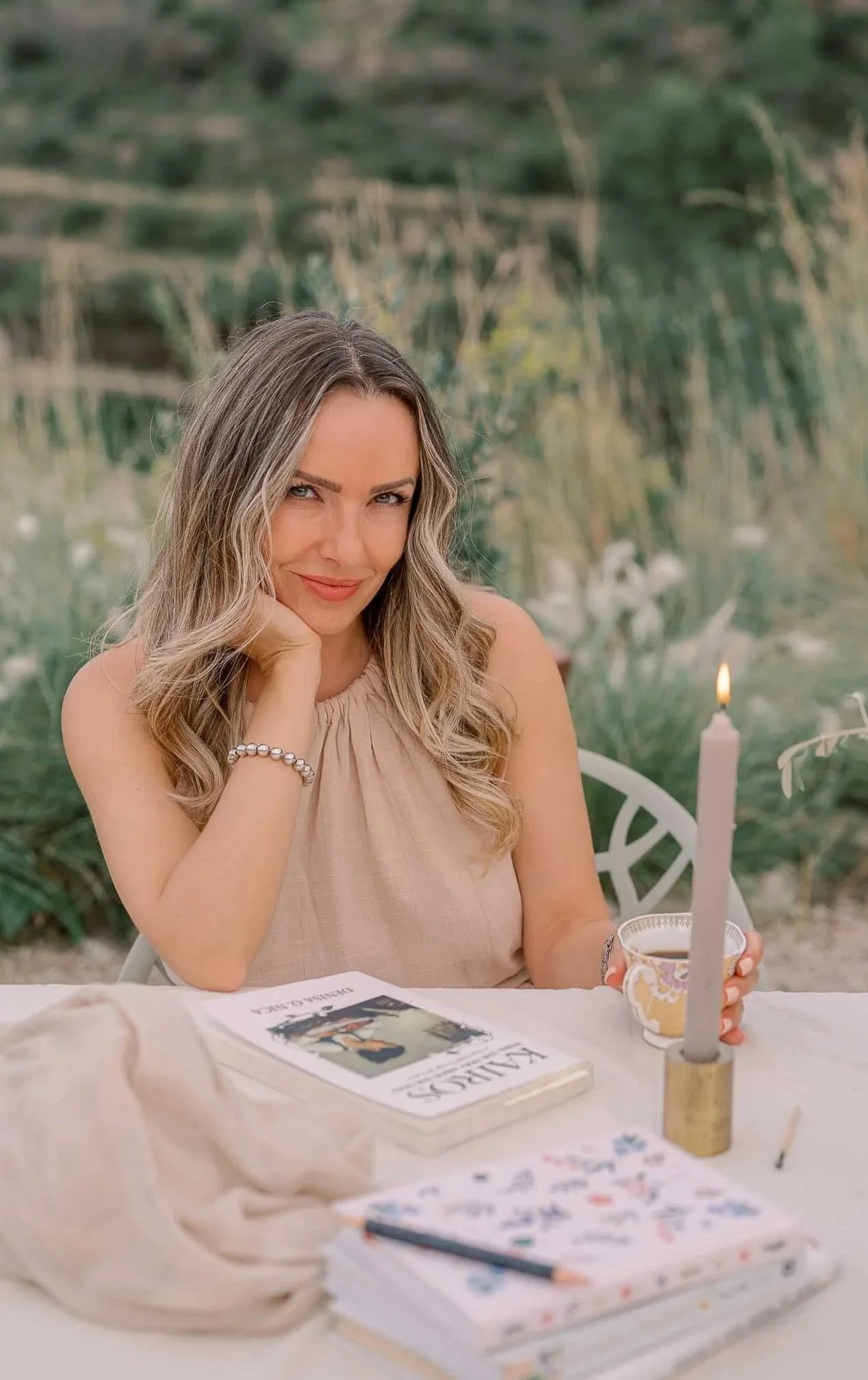 A woman with wavy blonde hair sitting at a table outdoors, smiling at the camera. On the table are a book titled "VIEWS" by Federico O. Mack, a notebook with colorful doodles, a lit pink candle in a gold holder, a floral-patterned teacup, and a soft beige cloth.