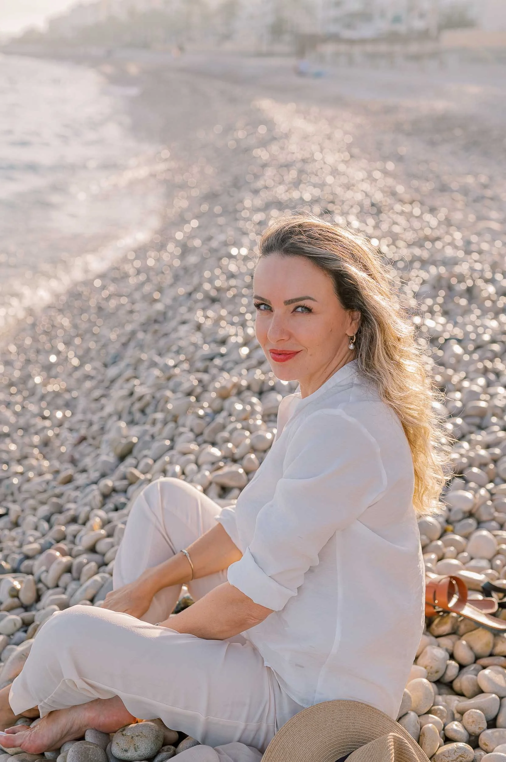 Denisa Nica sitting on a pebble beach with a slight smile, wearing a white outfit, with the sea in the background during sunset.