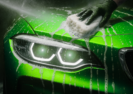 Person washing a bright green car with a sponge and soap, focusing on the car's headlight.