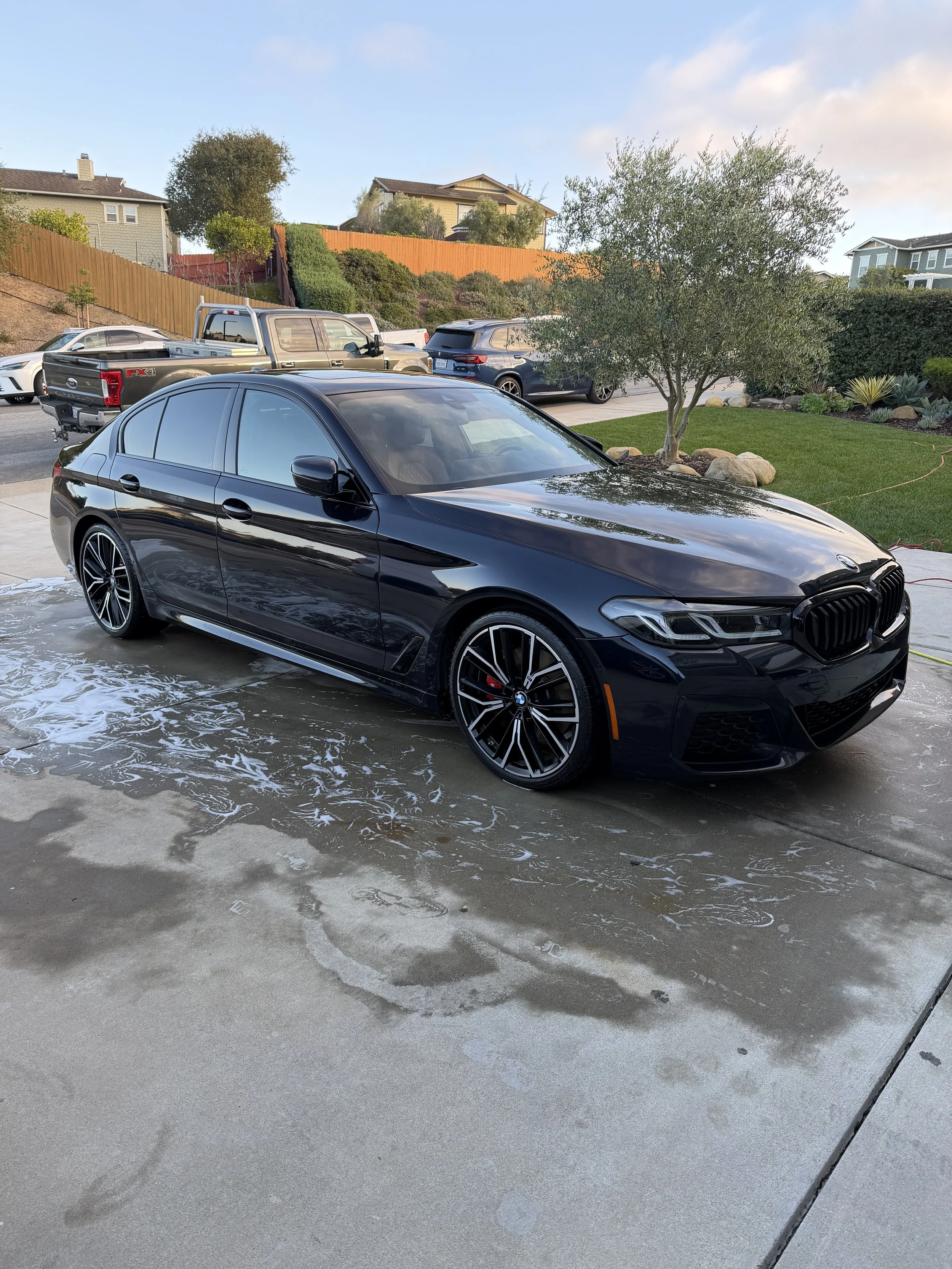 Black BMW sedan with foam and soap on the concrete driveway after washing, with water and soap suds visible.