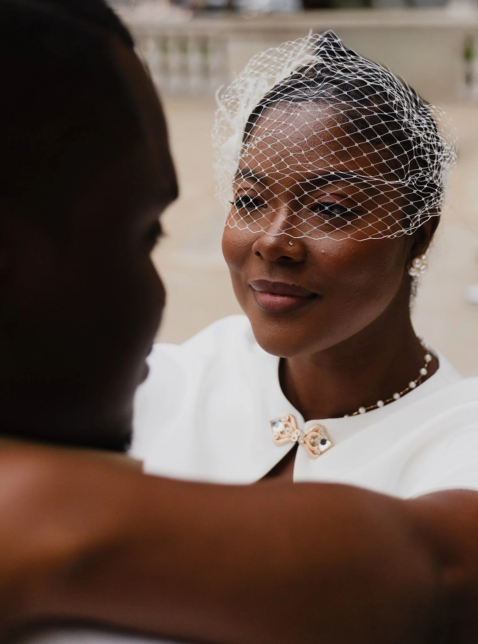 A woman wearing a birdcage veil and a white outfit is smiling during a wedding ceremony.