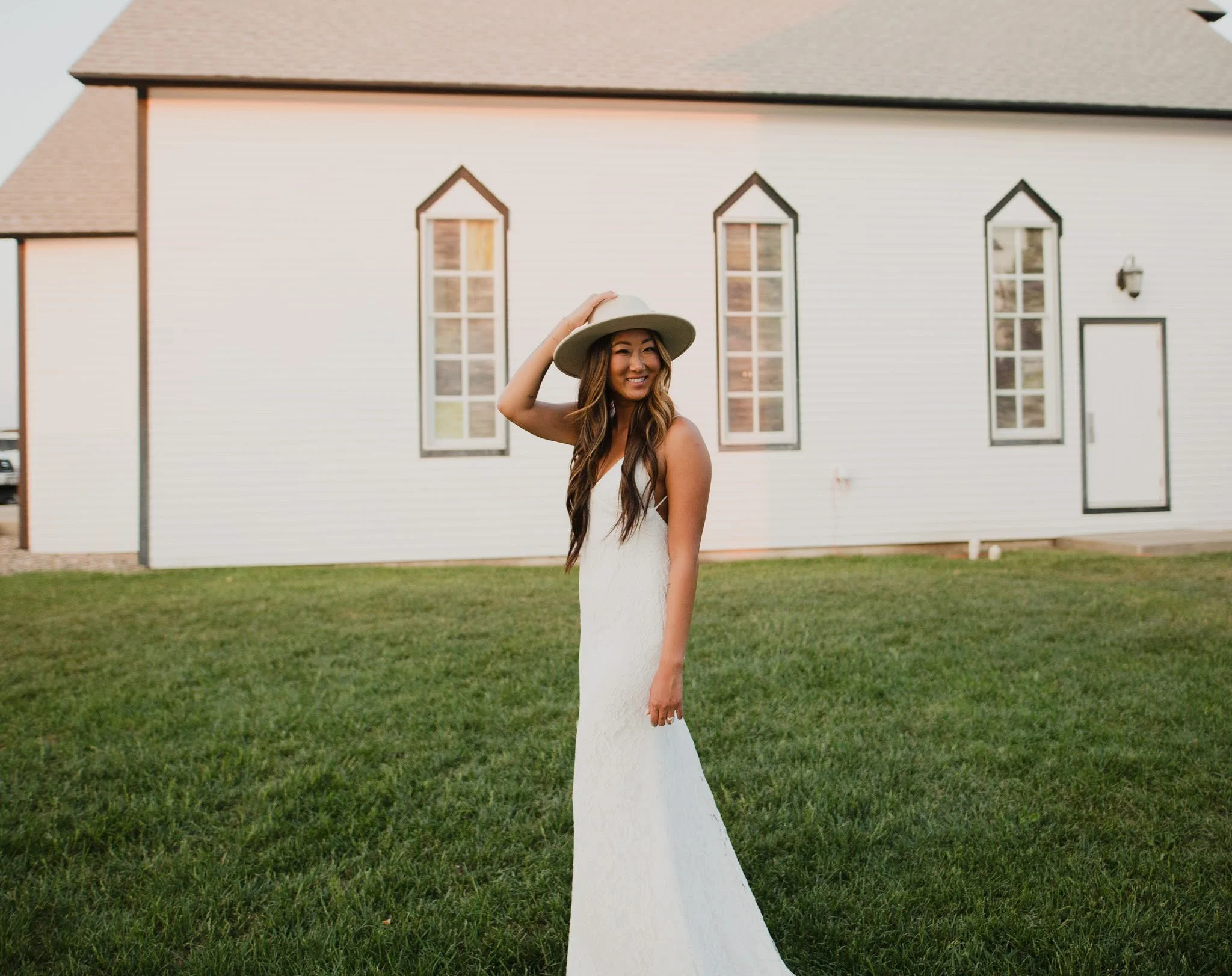 A woman in a white dress and wide-brimmed hat standing on a grassy lawn in front of a white building with three tall, narrow windows with cathedral-style arches, smiling and adjusting her hat.