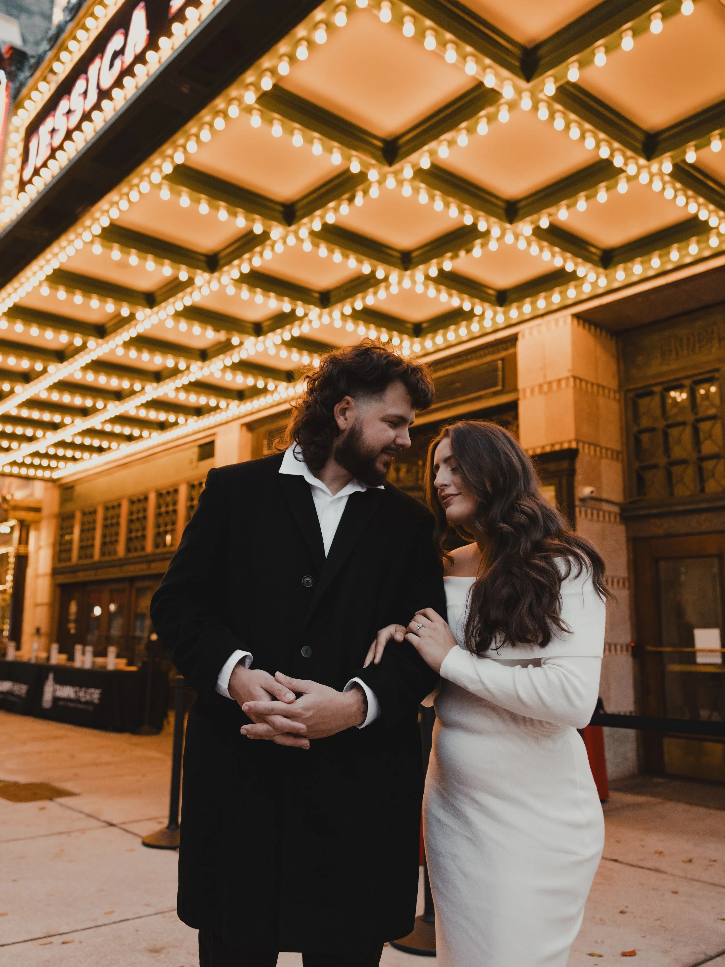 A romantic couple stands close under a decorated marquee with string lights. The man has long hair and a beard, wearing a black coat over a white shirt. The woman has long wavy hair, dressed in a white dress, holding his hand with both of hers. They 