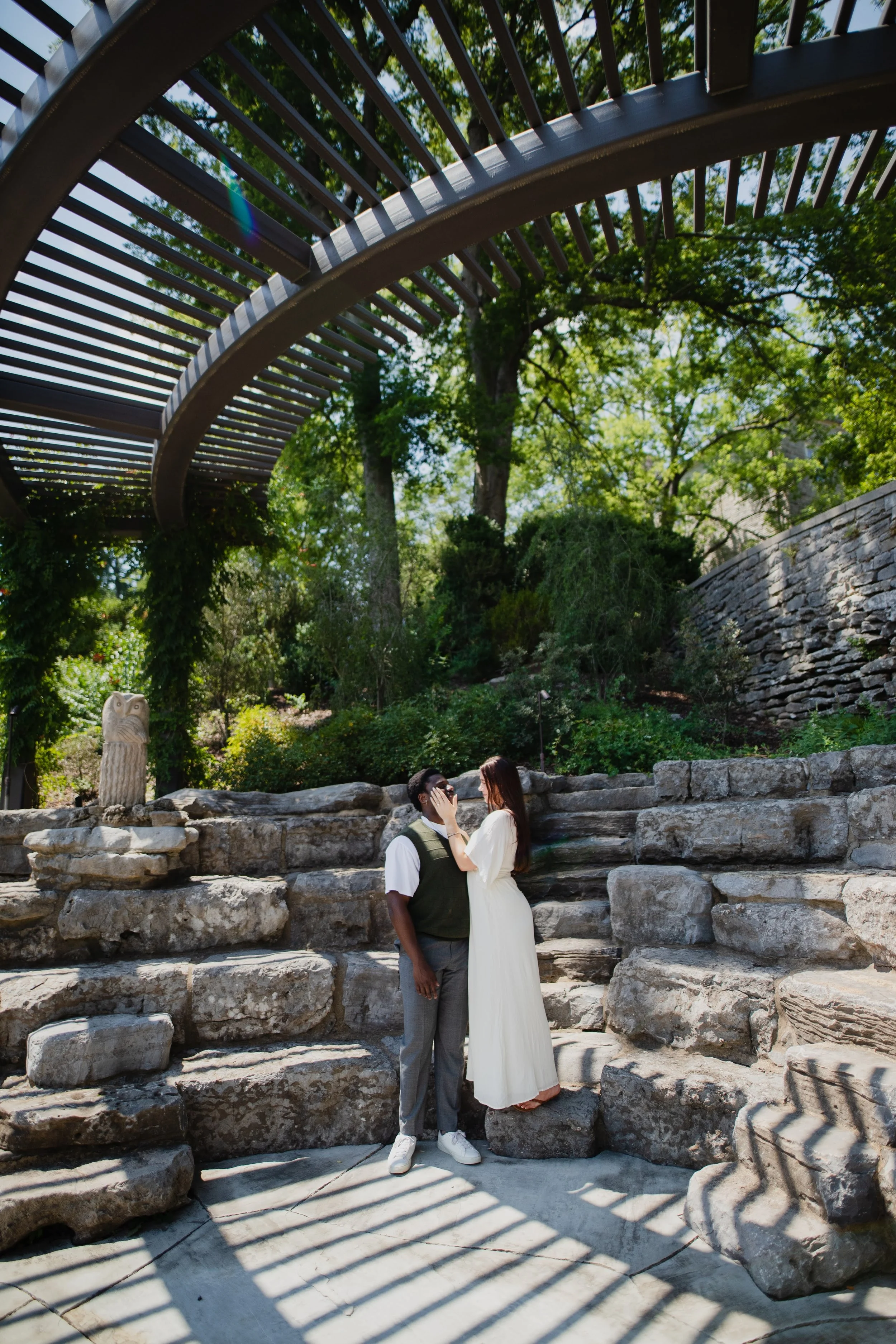 A couple standing on stone steps in a park-like setting, with the woman touching the man's face, both looking at each other affectionately, surrounded by trees and greenery, with a stone owl sculpture nearby and a modern metal pergola overhead.