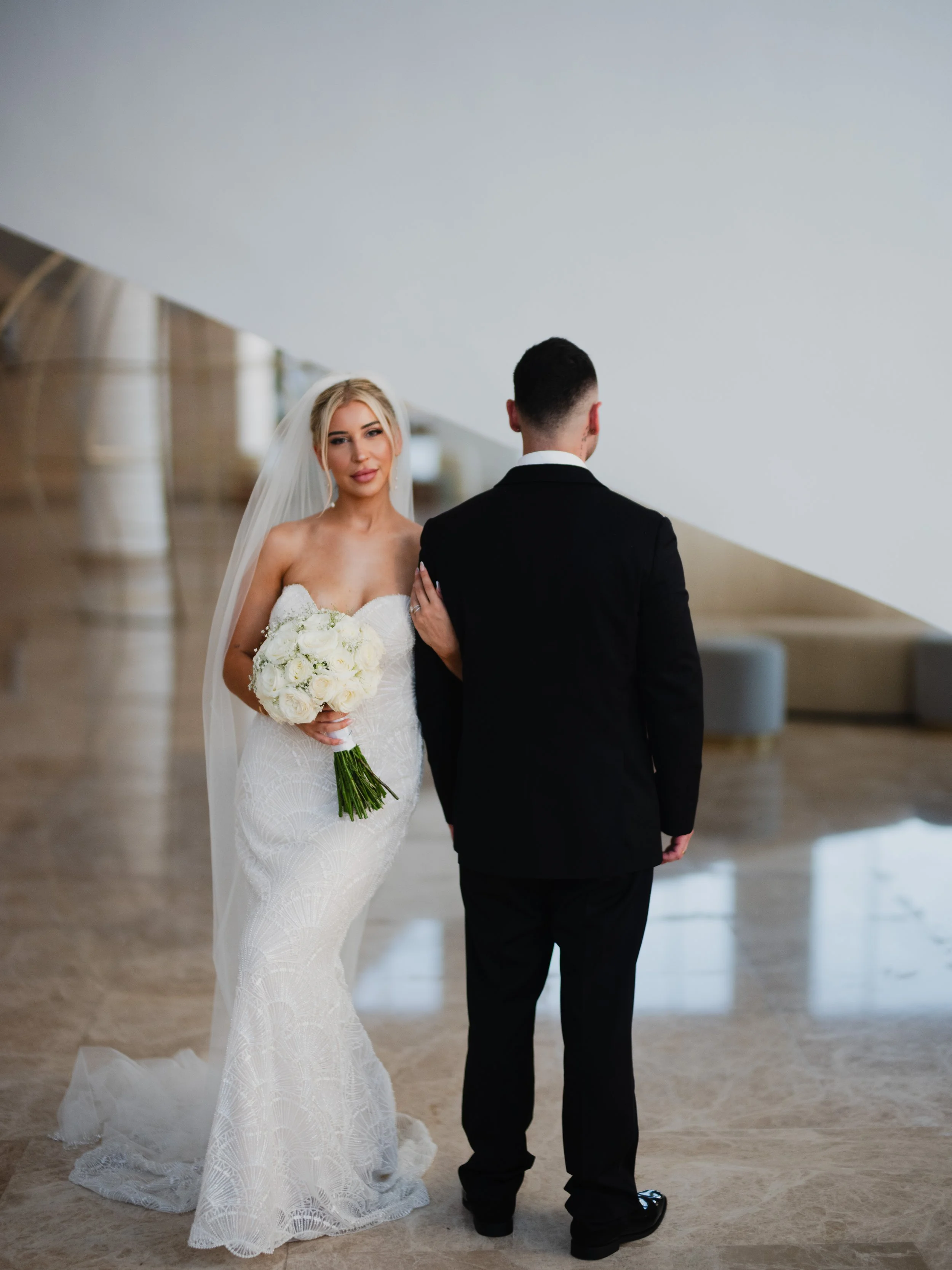 Bride holding a bouquet of white roses, standing next to groom in a black suit, inside a modern building.