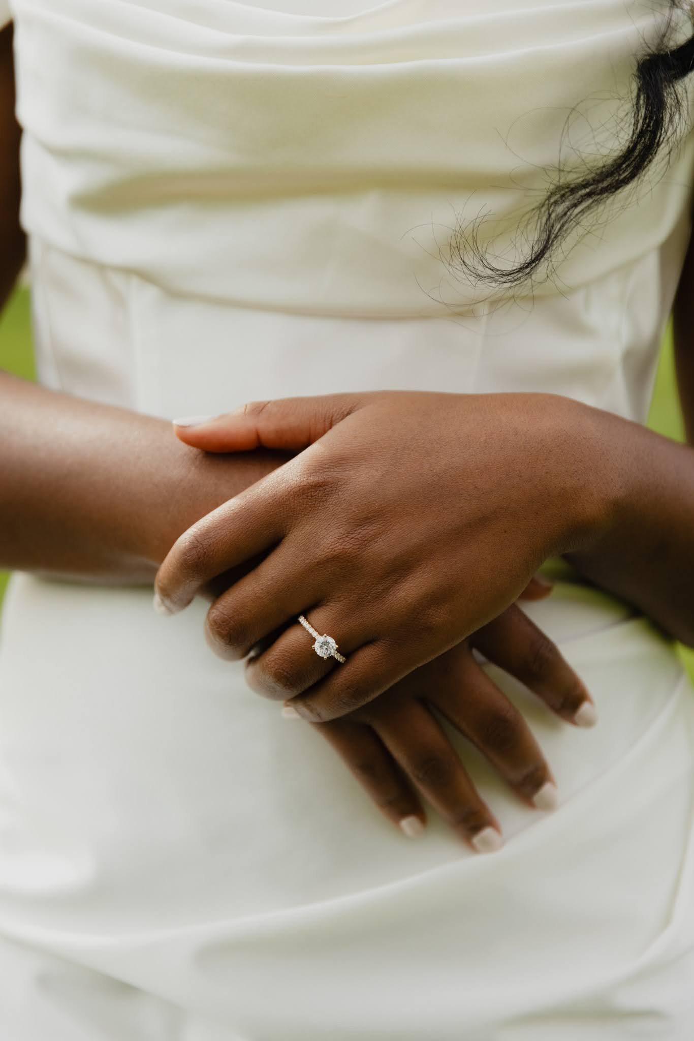 Close-up of a woman's hand with a diamond engagement ring, resting on her lap, with a white dress in the background.