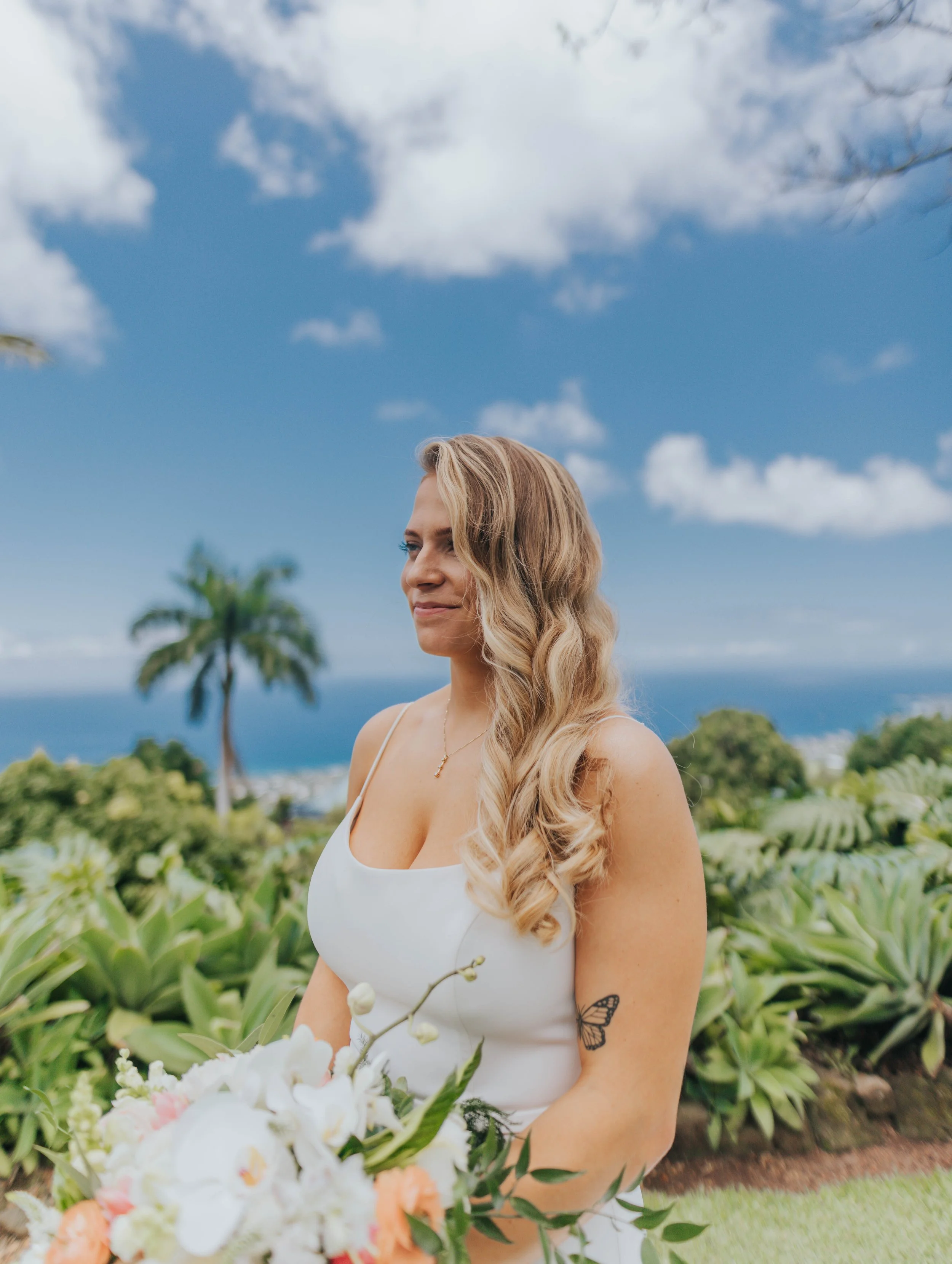 A woman with long, wavy blonde hair standing outdoors on a sunny day, holding a bouquet of flowers, with palm trees and the ocean in the background.