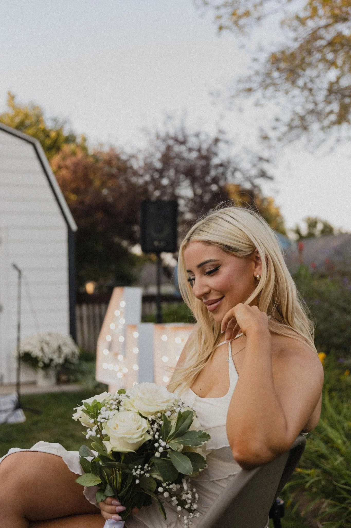 A woman with blonde hair sitting outdoors, holding a bouquet of white roses and greenery, smiling softly, with trees and a white shed in the background during sunset.