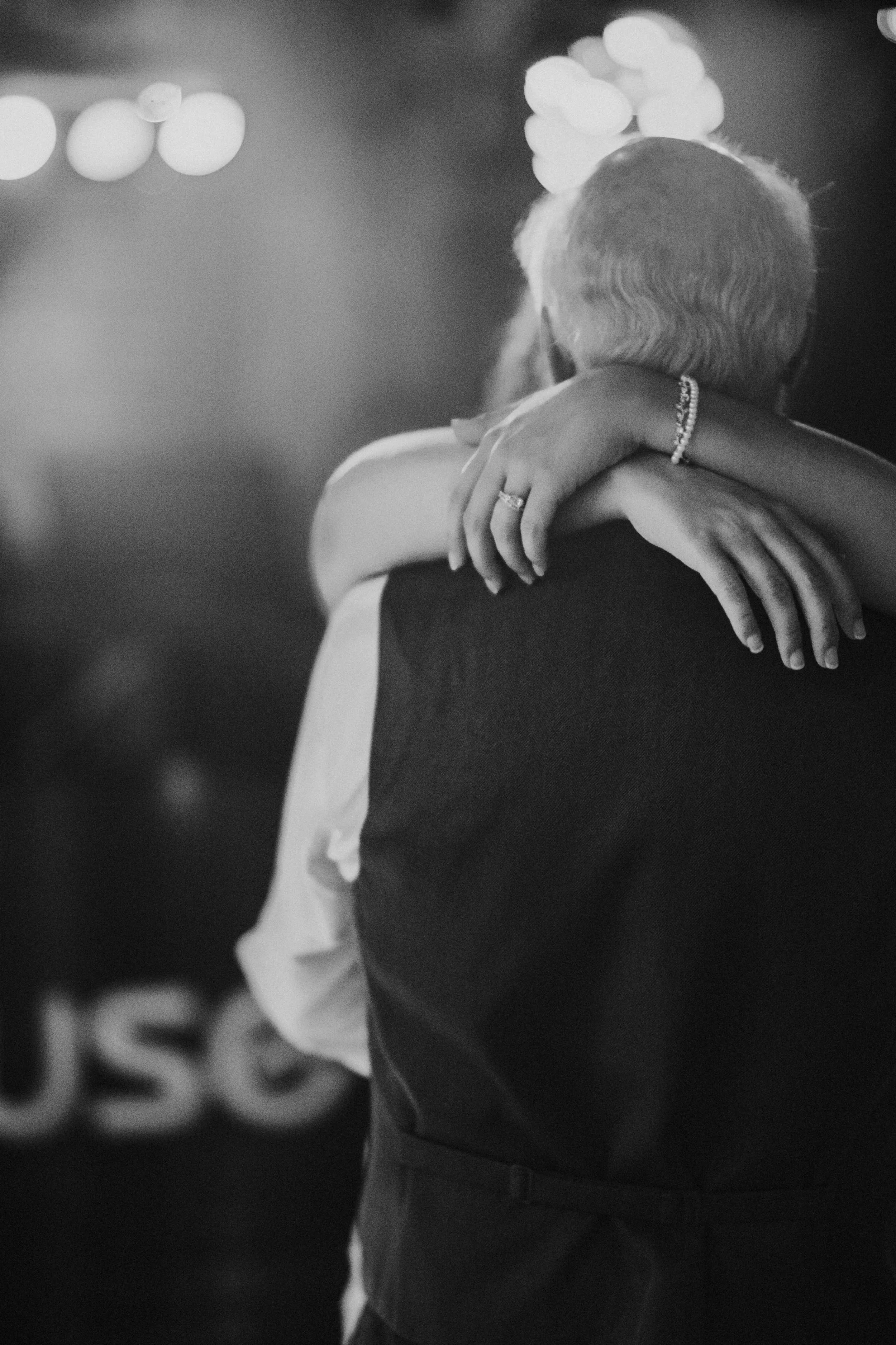 Black and white photo of a woman and a man embracing, with the woman's arms wrapped around the man's shoulders. The woman is wearing jewelry, including a ring and a bracelet. The photo is blurred in the background, with some lights visible.