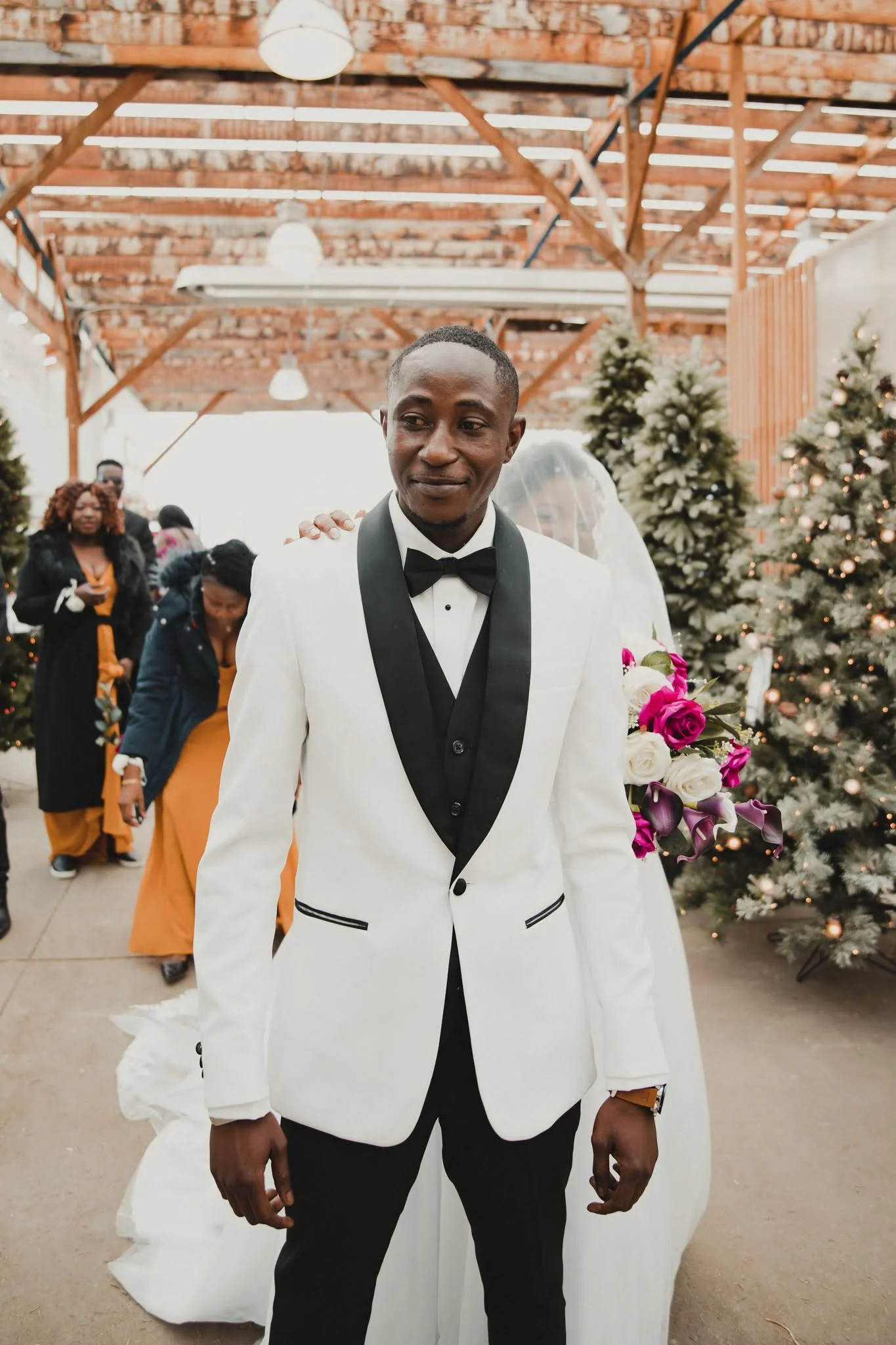 A groom in a white tuxedo with black lapel and black bow tie standing in front of a decorated Christmas tree, with wedding guests in the background.