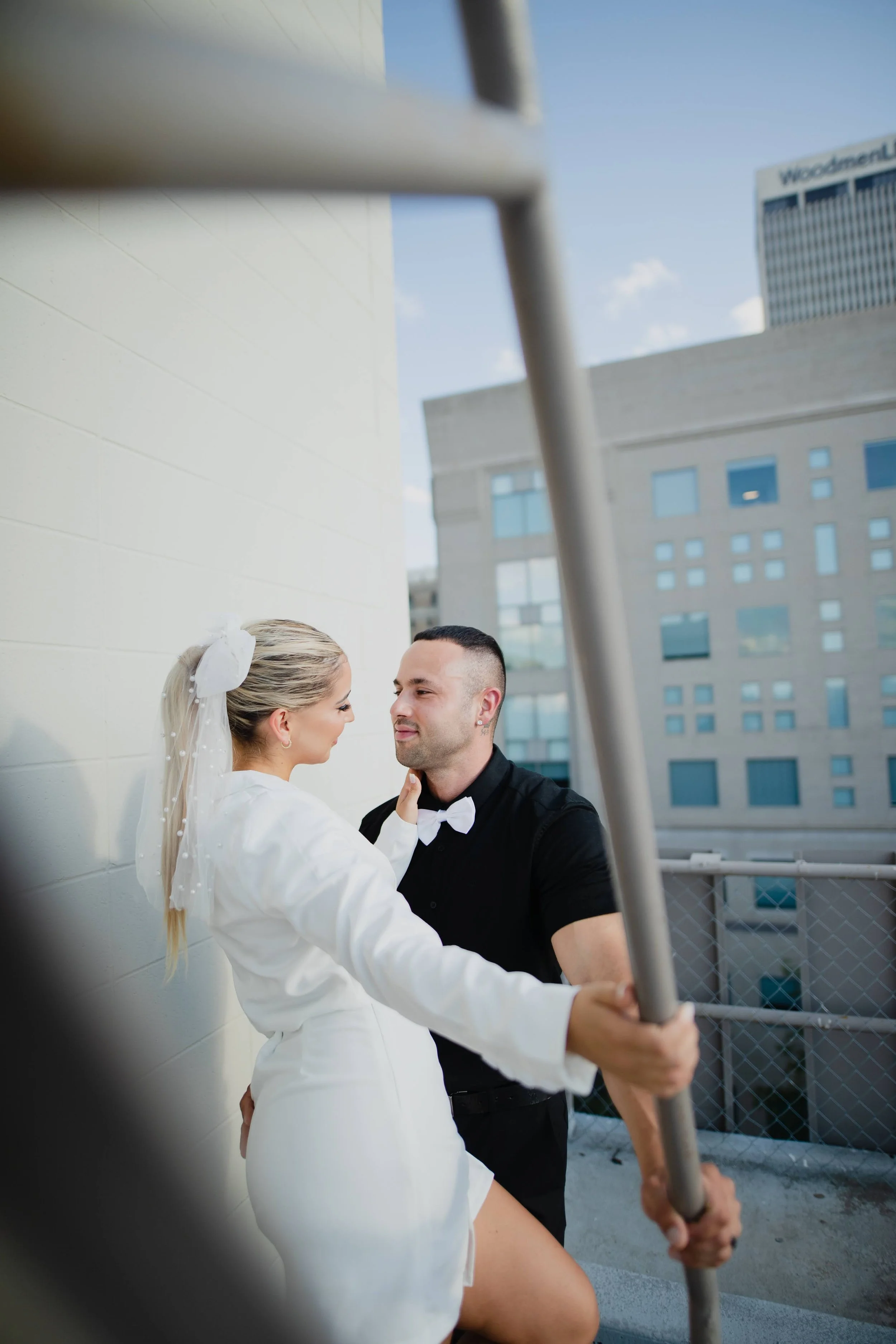 A woman in a white dress and a man in a black shirt with a white bow tie are looking at each other on a rooftop with city buildings in the background. She is holding onto a metal railing.