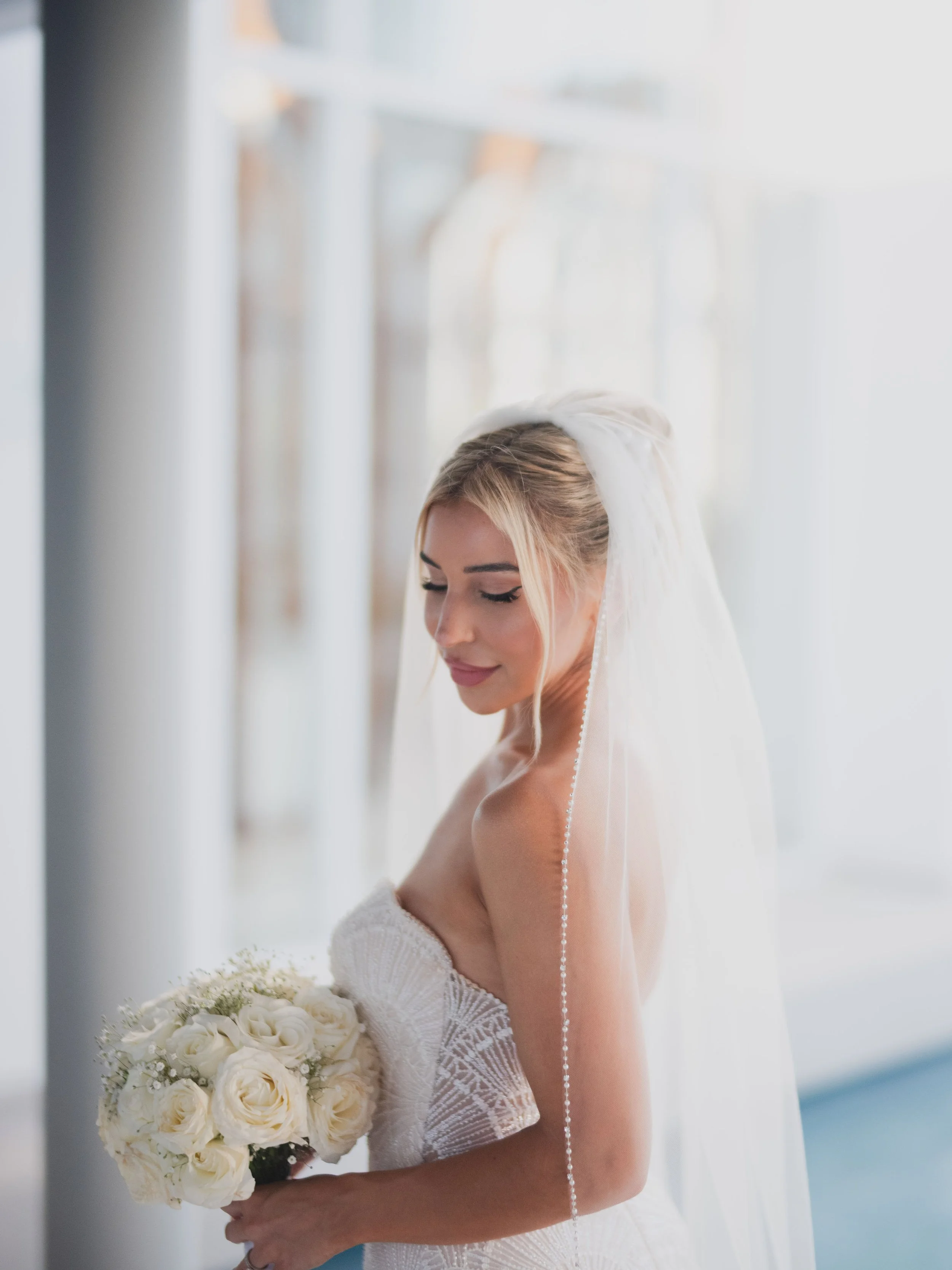 A bride in a strapless wedding gown holding a bouquet of white roses, with a long veil, standing near large windows with natural light.
