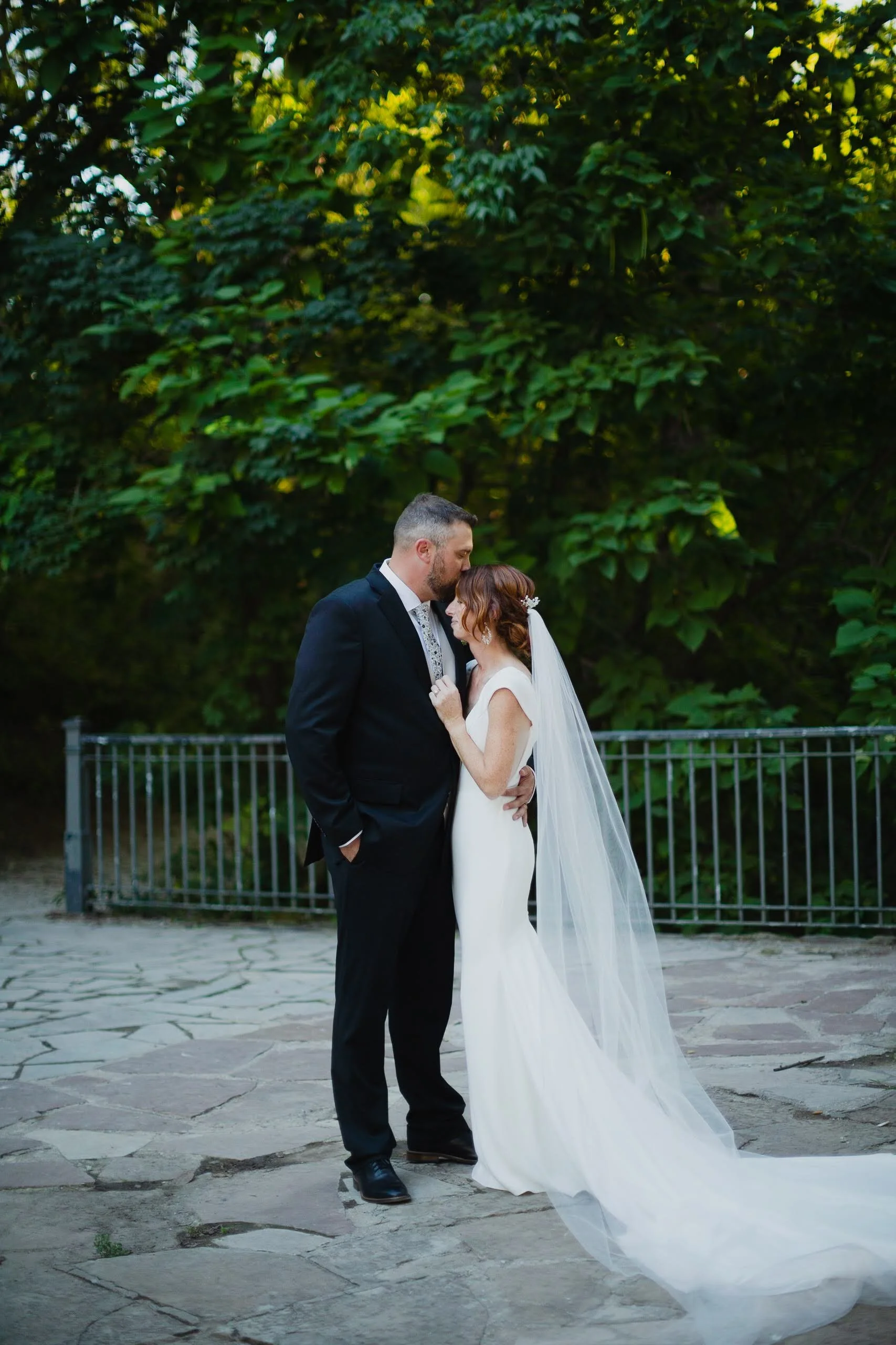A bride and groom in wedding attire standing close, with their foreheads touching, outdoors surrounded by green foliage, on a stone-paved area.