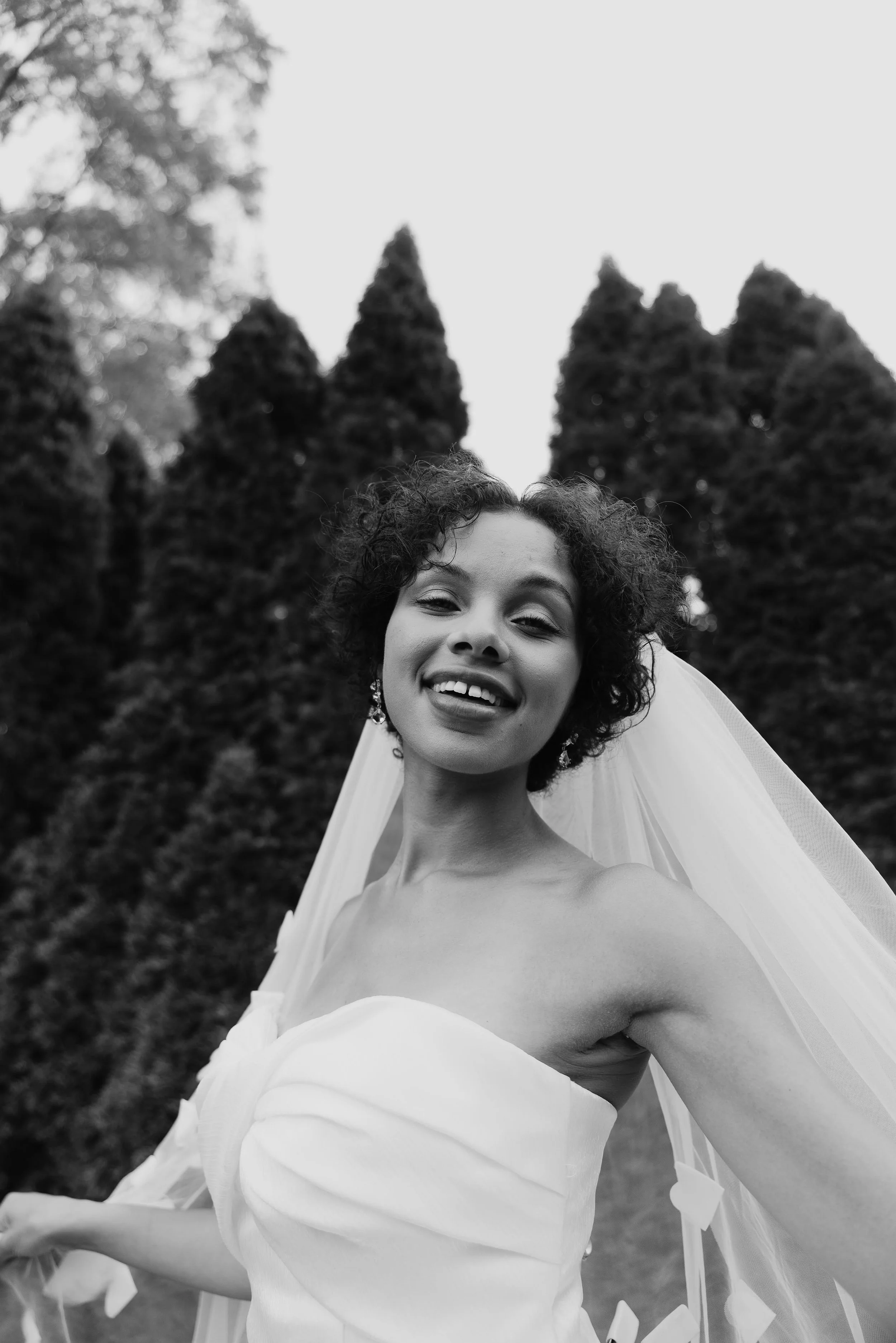Black and white photo of a woman smiling in a wedding dress and veil outdoors with tall trees in the background.