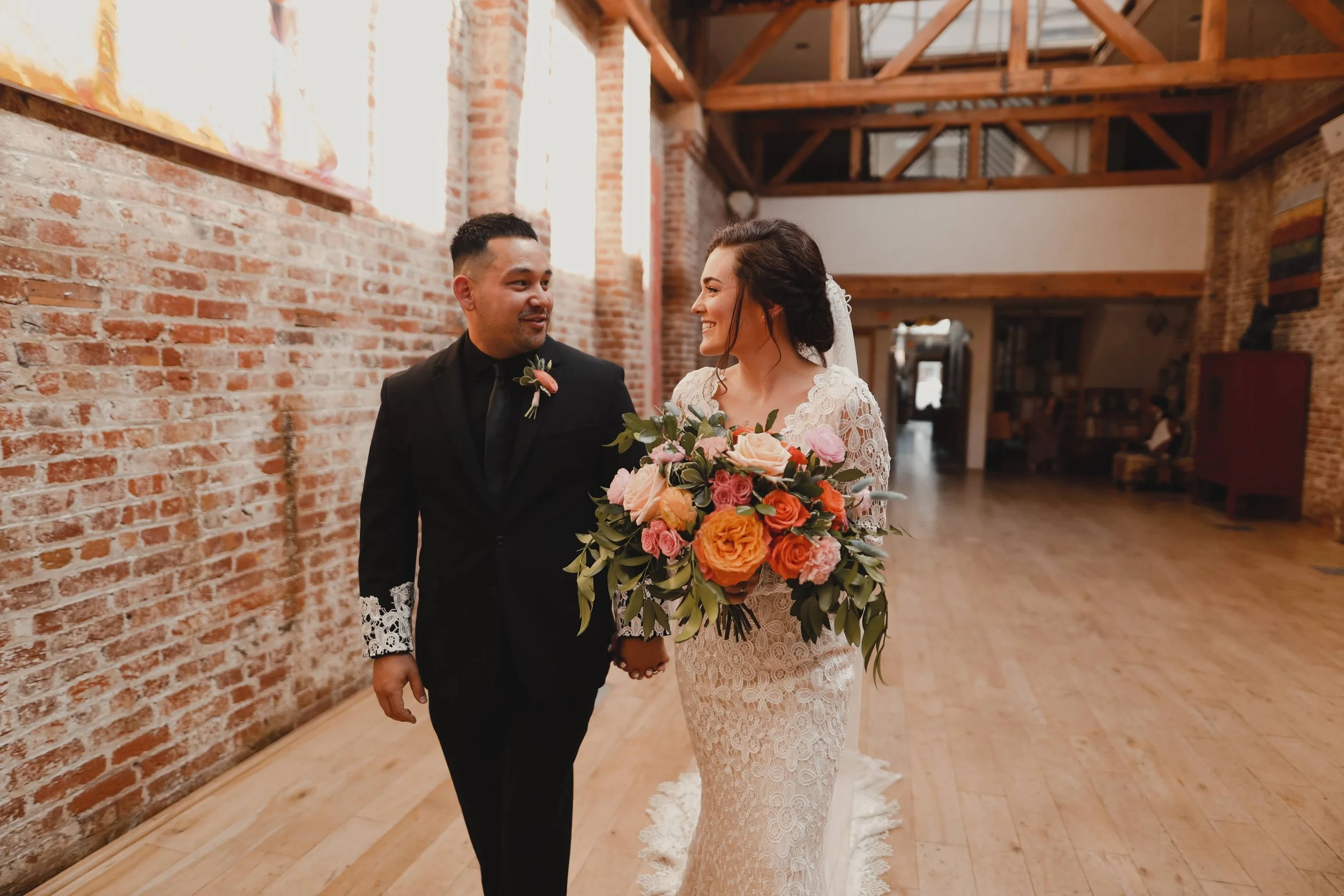 A bride and groom holding hands and walking together in a rustic, brick-walled venue. The bride is wearing a lace wedding dress and holding a large bouquet of pink, orange, and peach flowers. The groom is dressed in a black suit with lace-cuffed sleeves and a boutonniere.