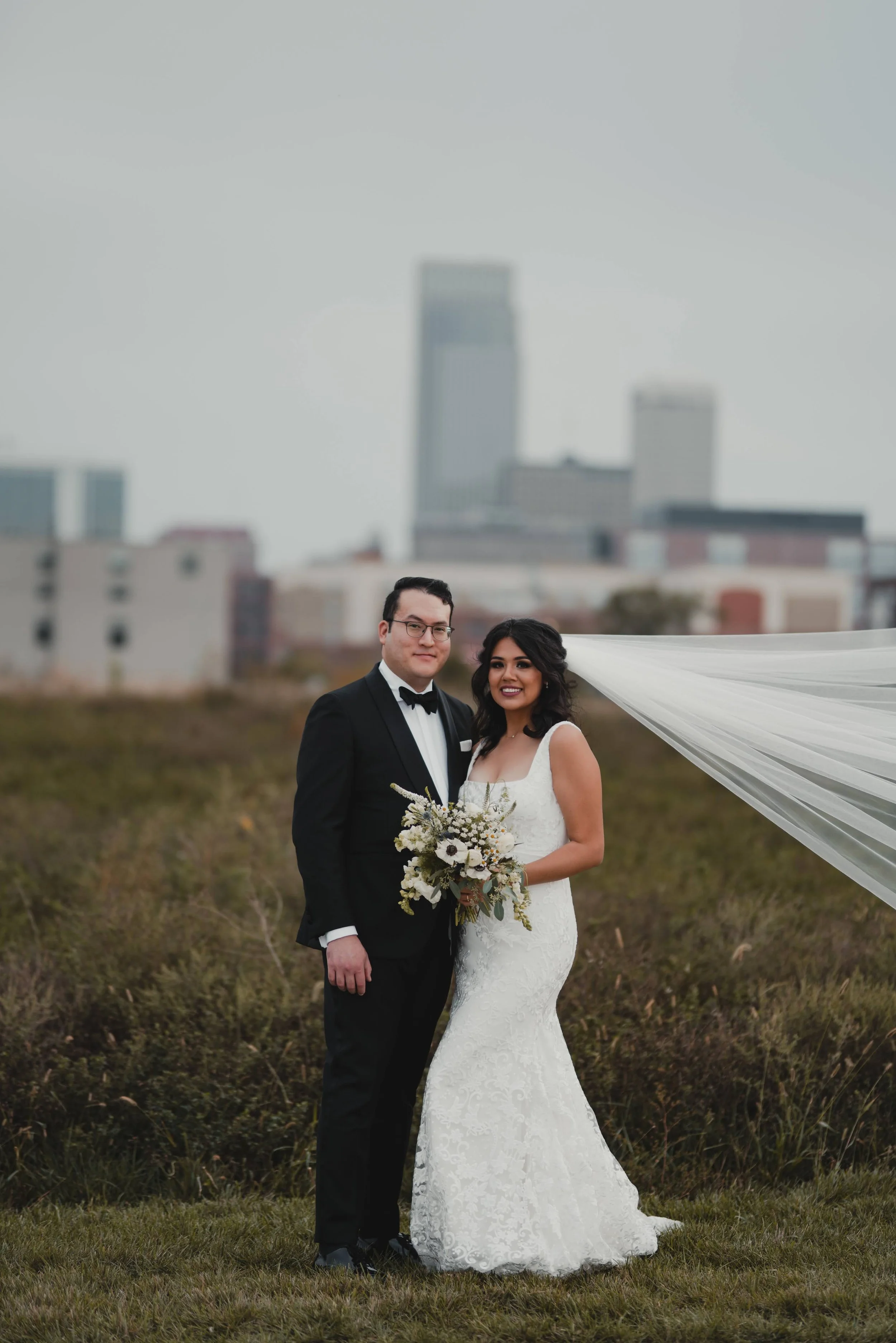 A newlywed couple standing outdoors in a field with city buildings in the background. The bride is wearing a white lace wedding gown and holding a bouquet, and the groom is dressed in a black tuxedo with a bowtie.