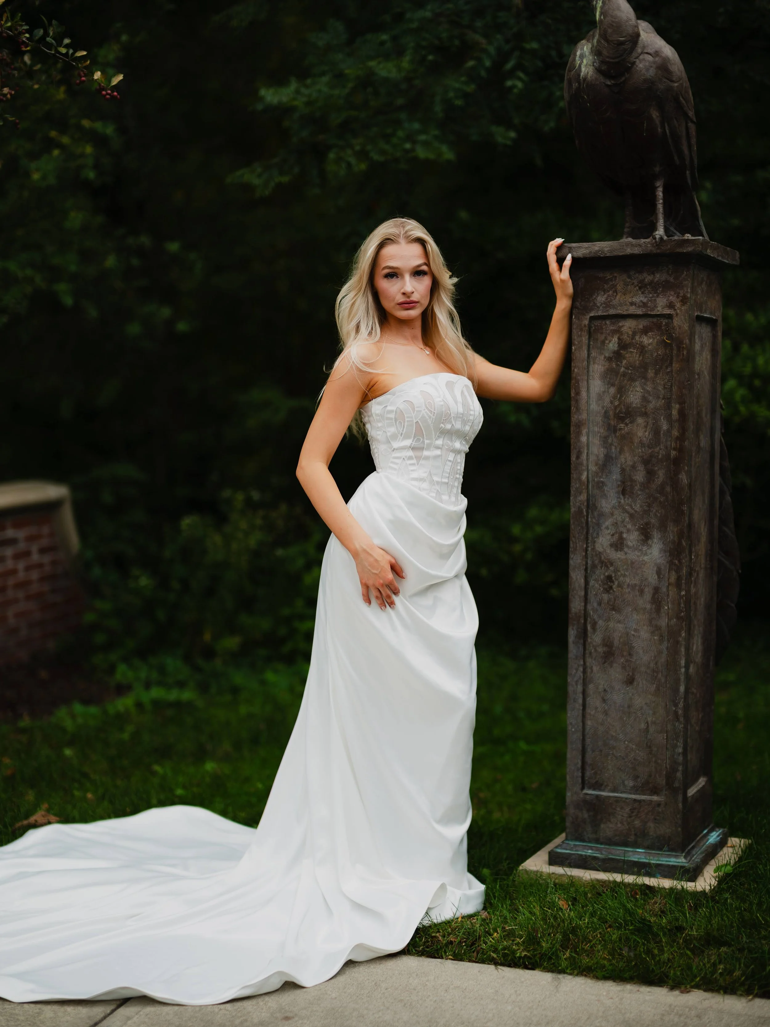 A woman in a white wedding dress standing outdoors next to a statue of a bird on a pedestal.