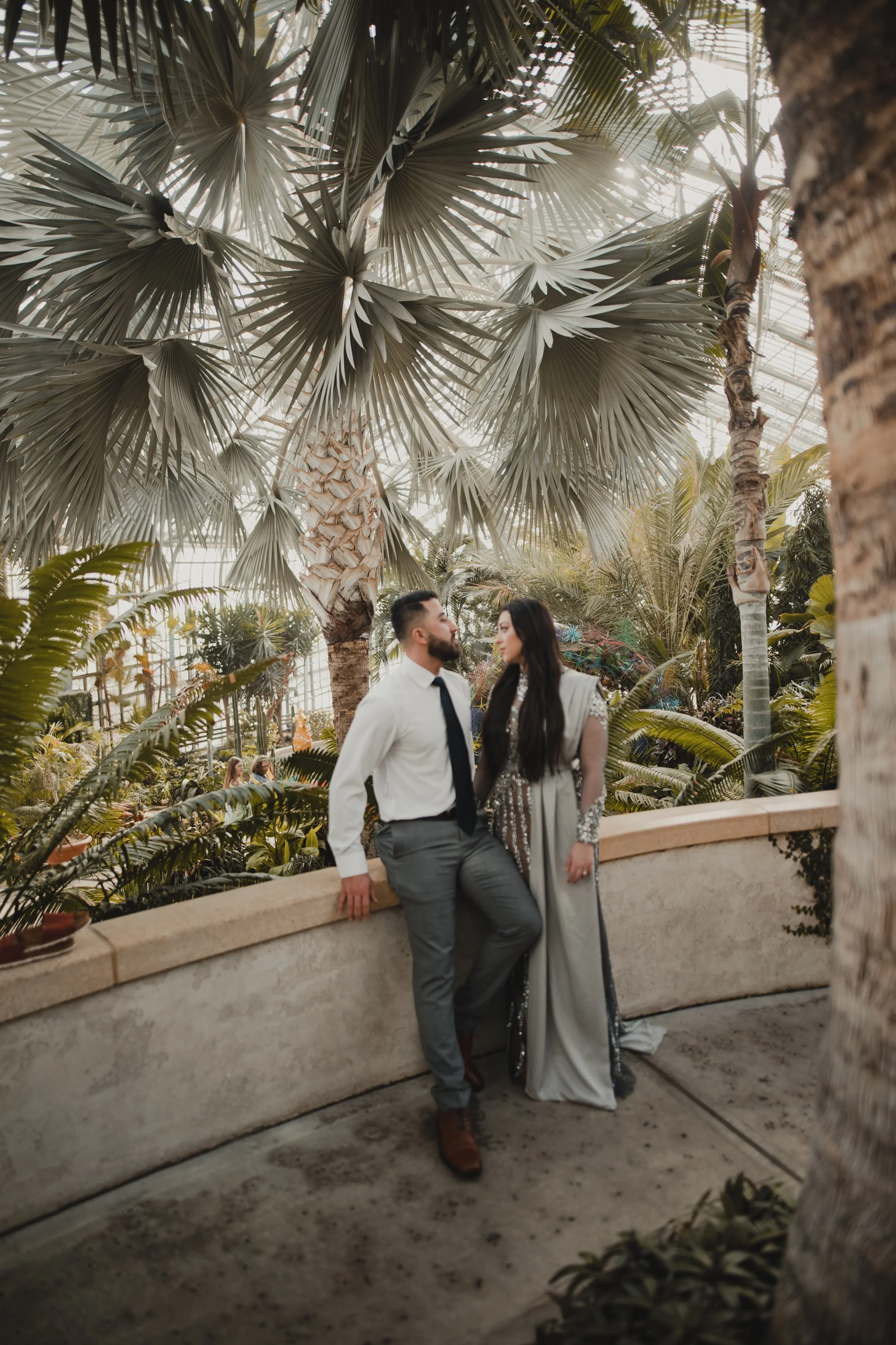 A couple in formal attire standing and sitting near a low wall in a tropical greenhouse with large palm leaves and lush greenery.
