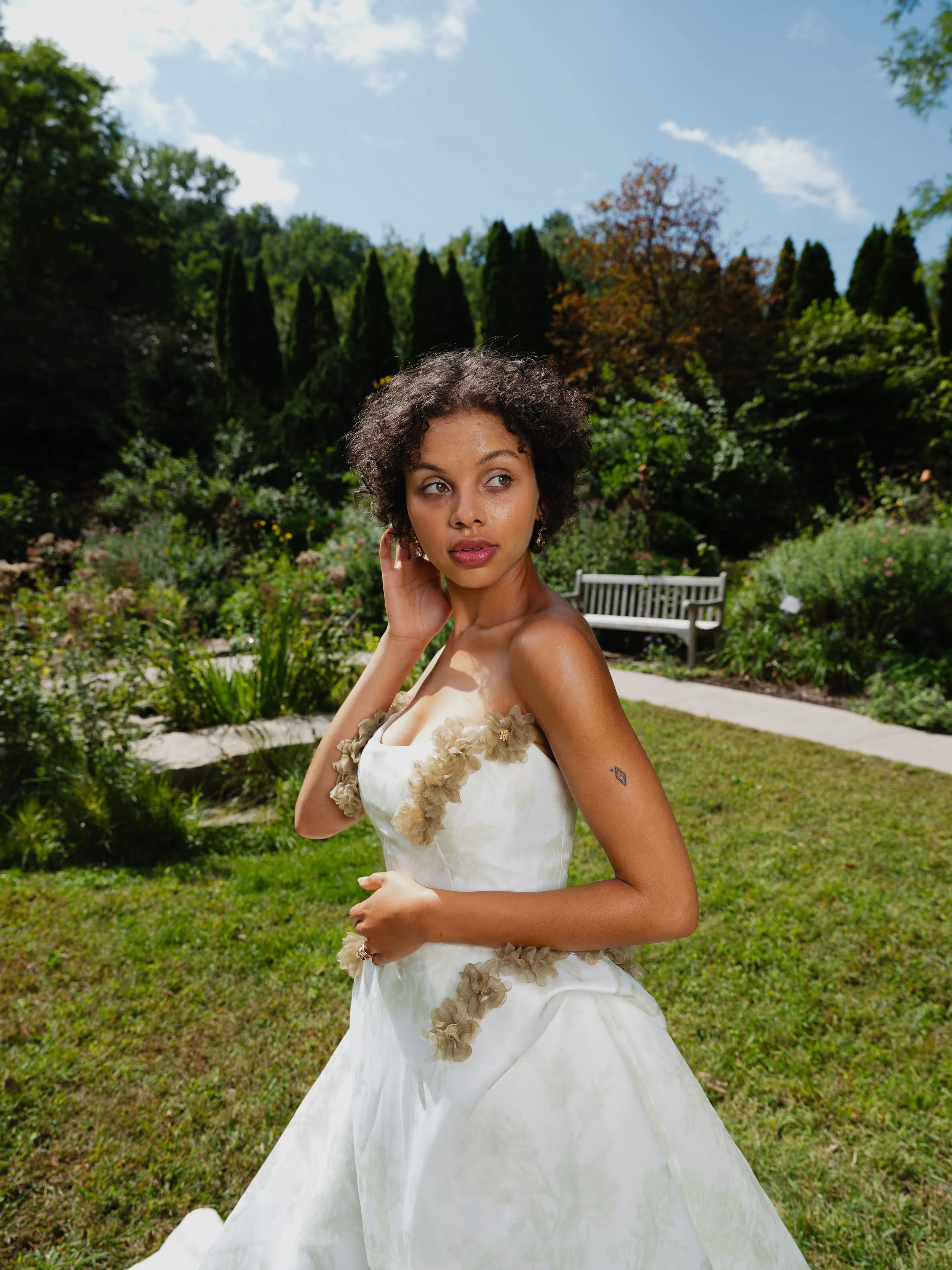 A woman in a white wedding dress with floral embellishments, standing on a lawn in a garden with trees and bushes, under a blue sky with some clouds.
