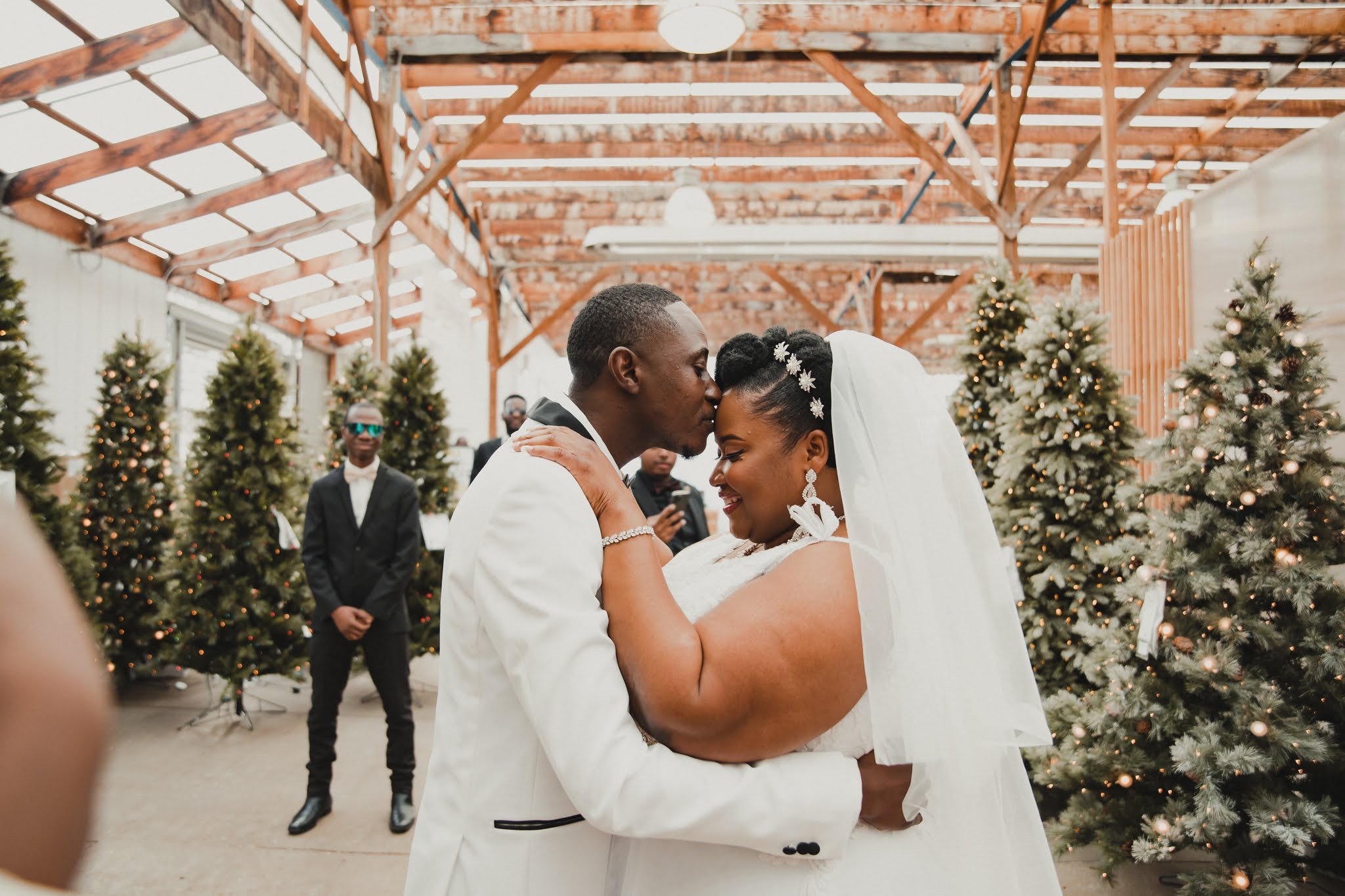A wedding couple shares a tender moment, with the groom kissing the bride on her forehead in a decorated venue with Christmas trees.