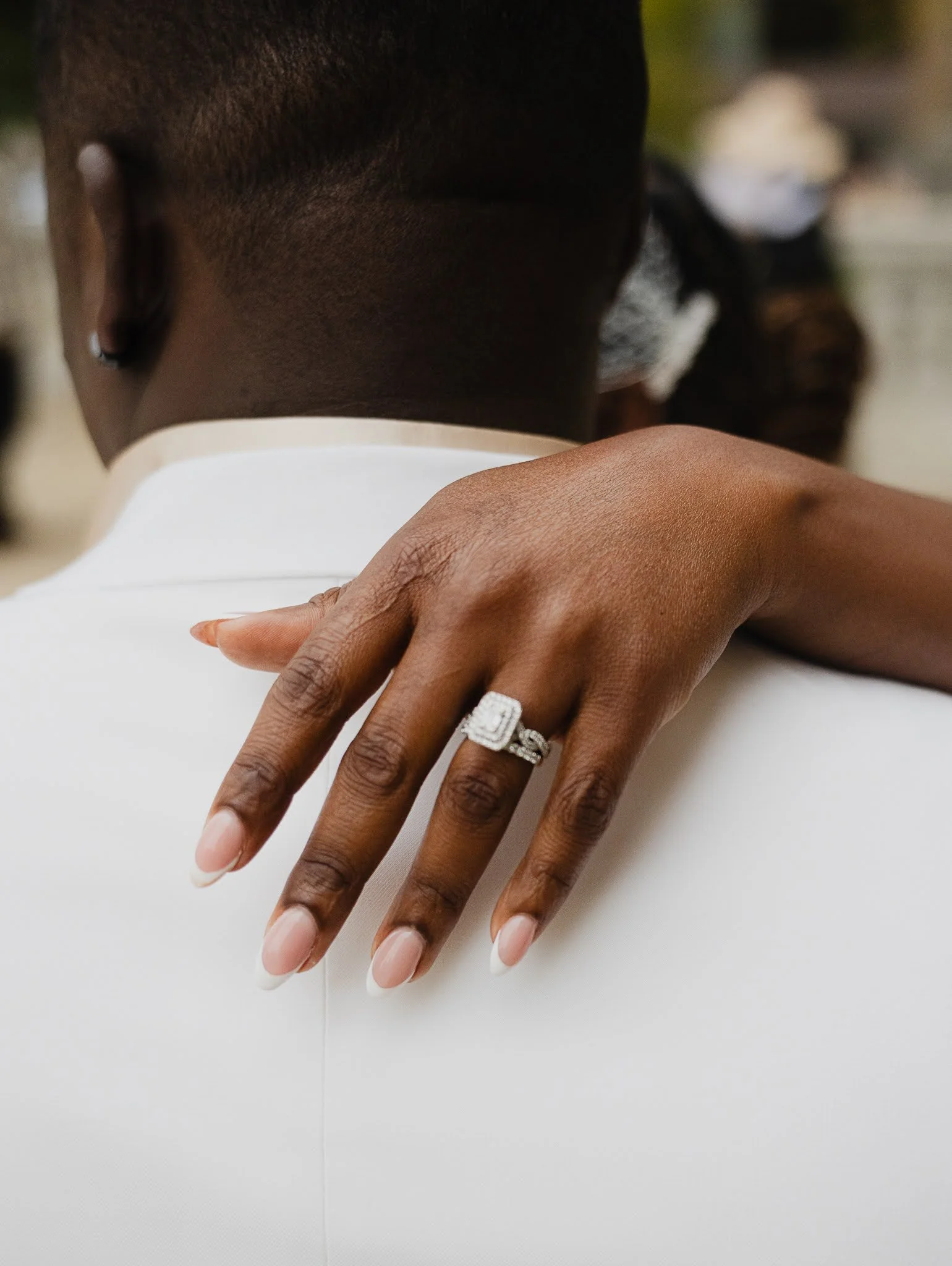 A woman with dark skin and a silver earring, resting her hand on a man's shoulder. She is wearing a large diamond ring on her finger, and her nails are manicured with a light pink polish. The man has short hair and is wearing a white shirt.