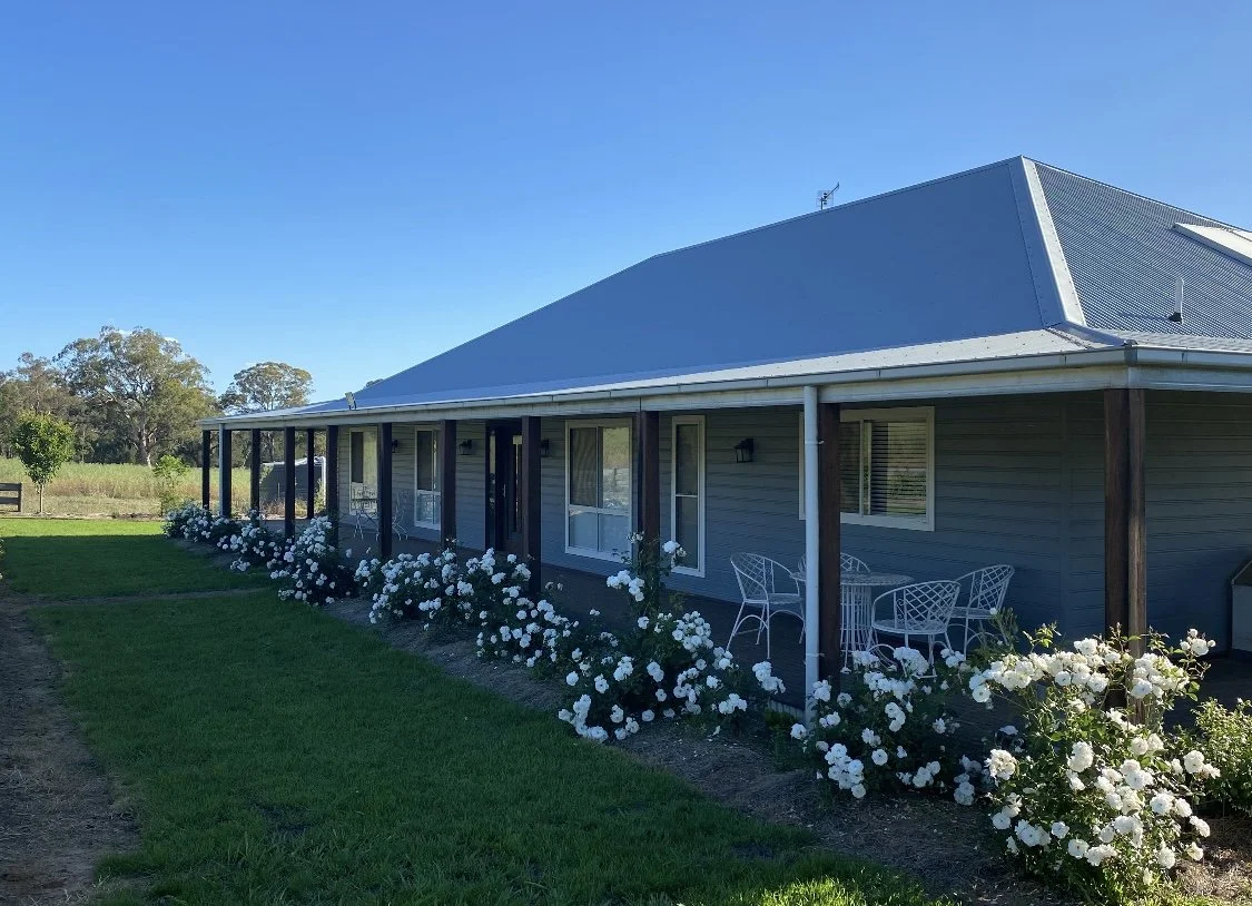 A residential house with a porch, surrounded by green grass and white flowering bushes, under a clear blue sky.