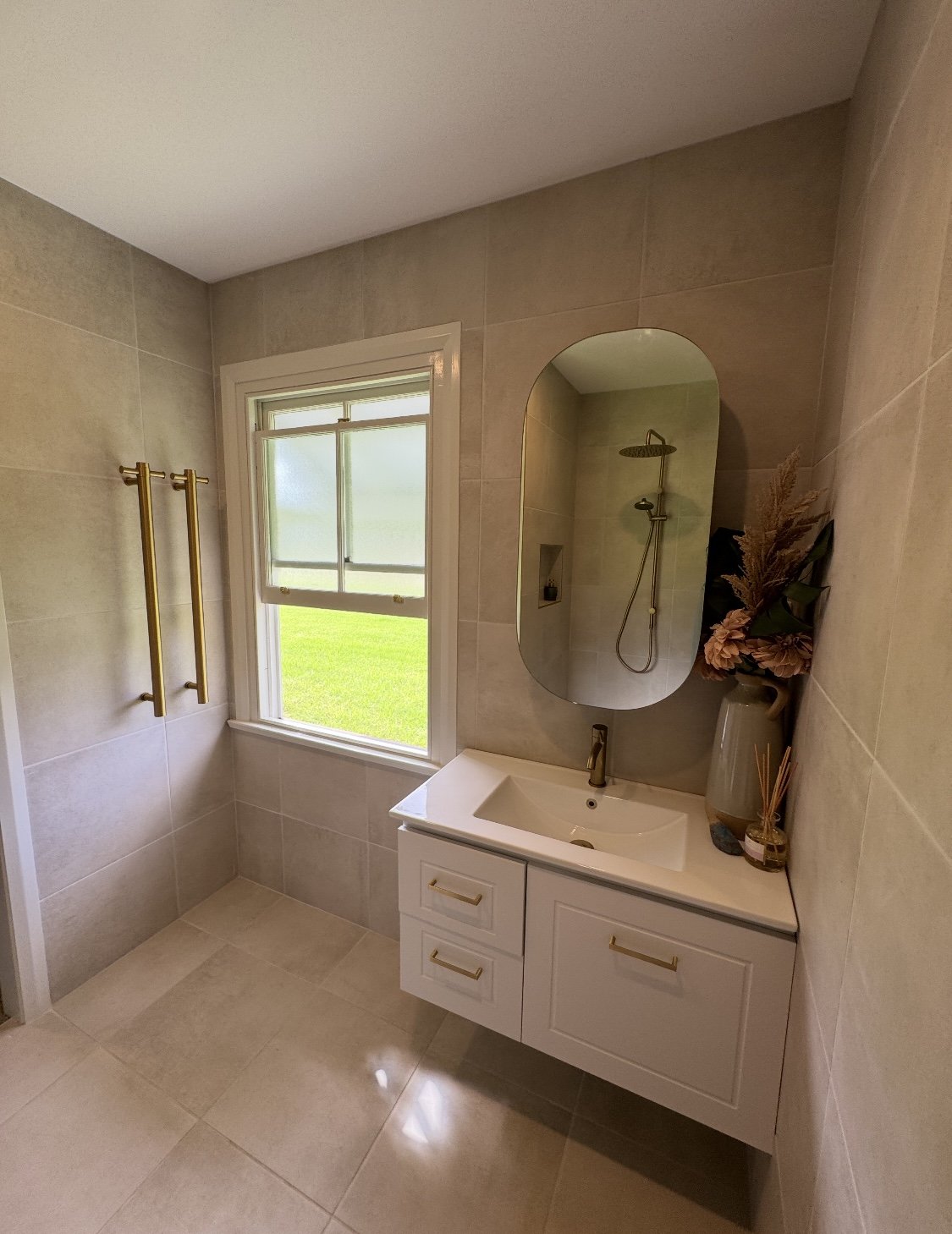 Modern bathroom with a window, a white vanity with gold handles, a large mirror, a vase with dried flowers, and a shower area.