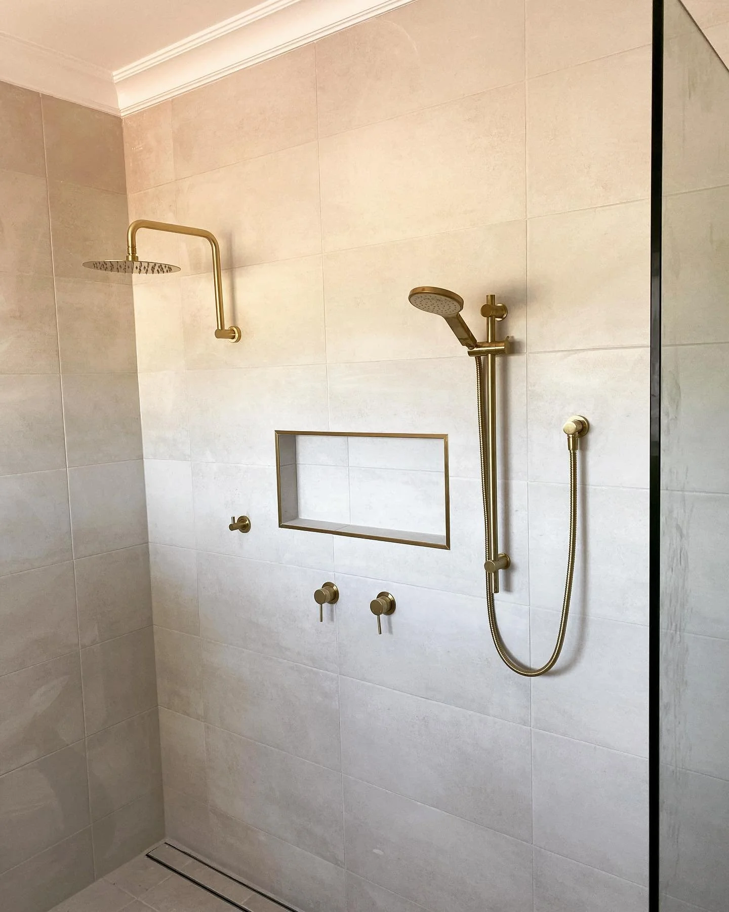 A modern shower with a gold showerhead, a handheld spray, and a built-in niche in a beige tiled wall.