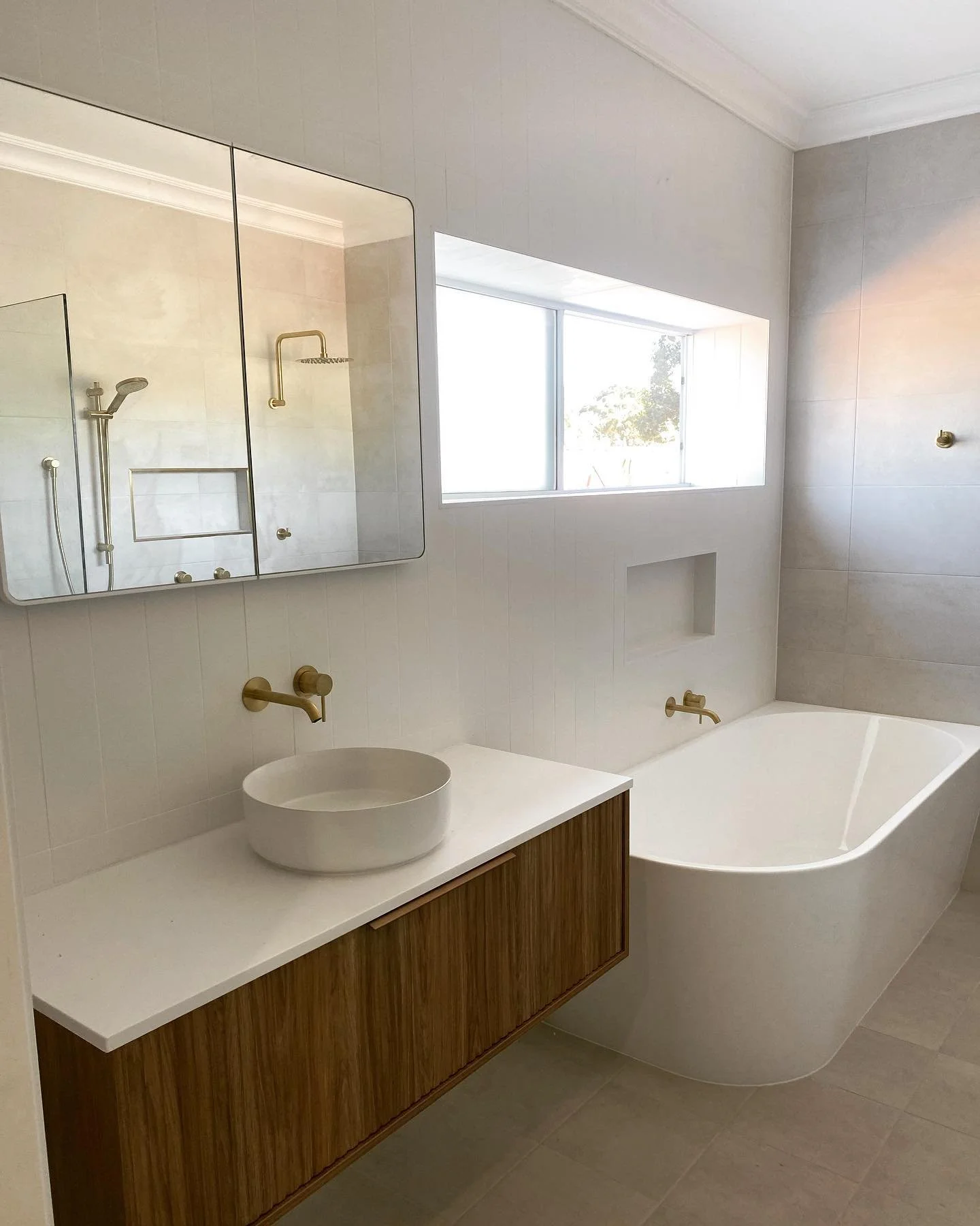 Modern bathroom with a white bathtub, a white round vessel sink on a wooden vanity, and a large mirror above the sink, with a window allowing natural light.