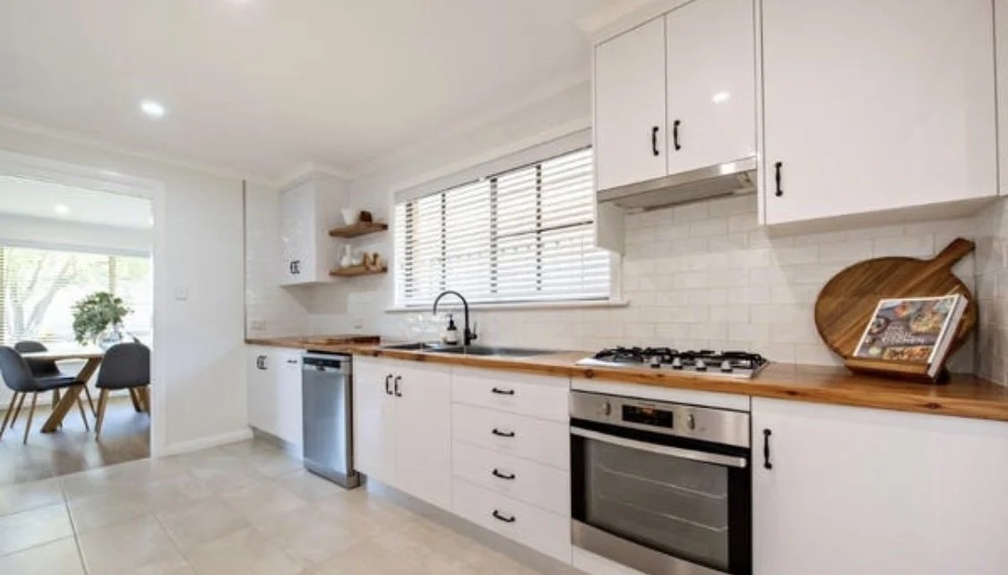 Modern kitchen with white cabinets, wooden countertops, stainless steel appliances, a window with blinds, and a view into a dining area with a table and chairs.