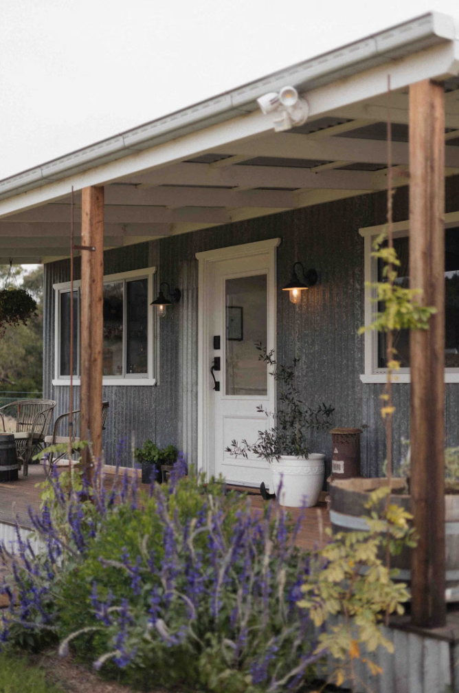 Front porch of a house with outdoor chairs, potted plants, and purple flowers in the foreground.