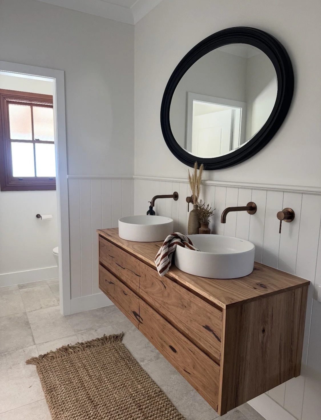Modern bathroom with a wooden vanity with two vessel sinks, a round mirror above, and a window with natural light. The bathroom features beige tiled floors and white wainscoting.