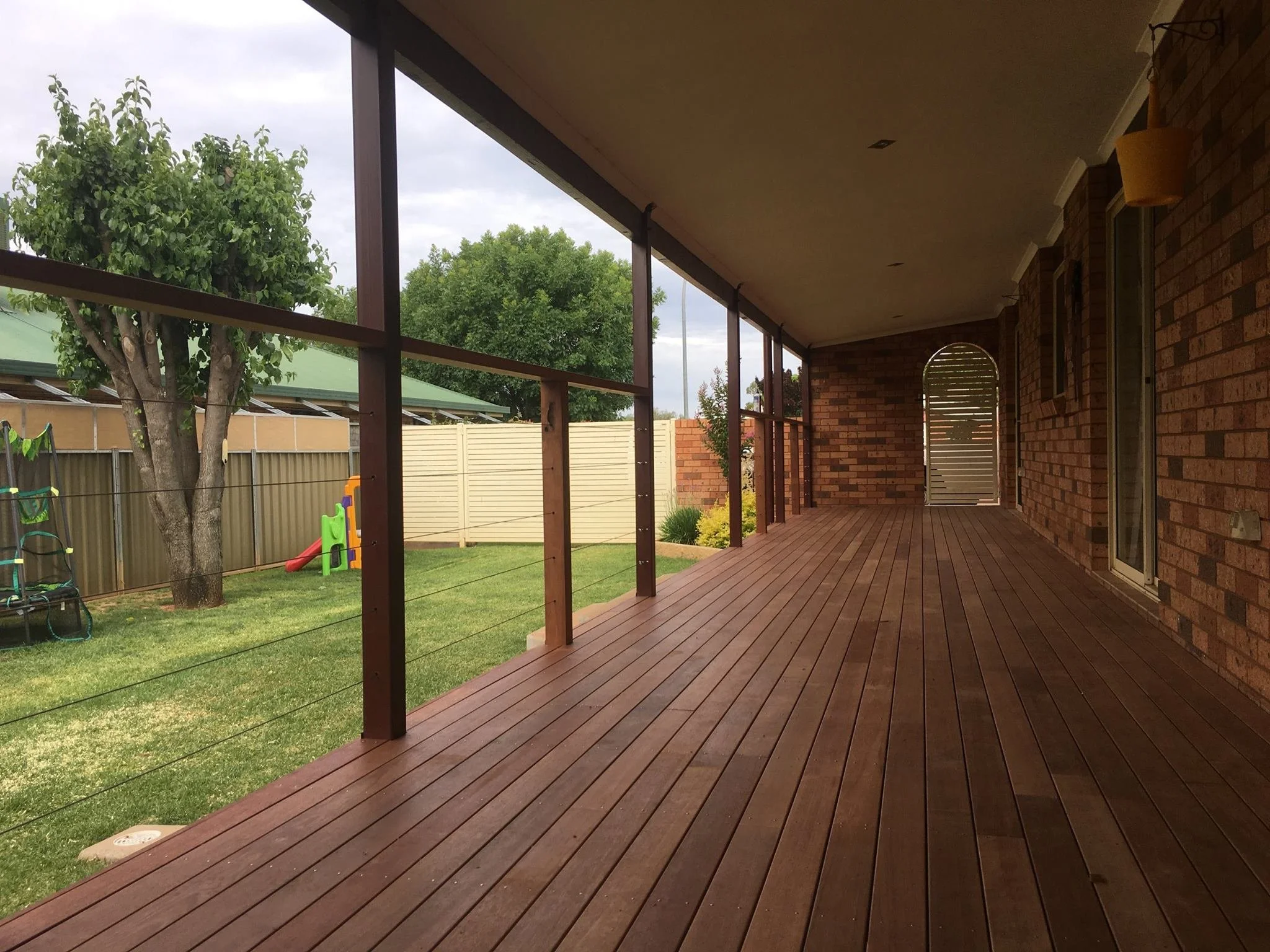 Empty wooden porch with a railing overlooking a backyard with grass, trees, a slide, and a fence, under an overcast sky.