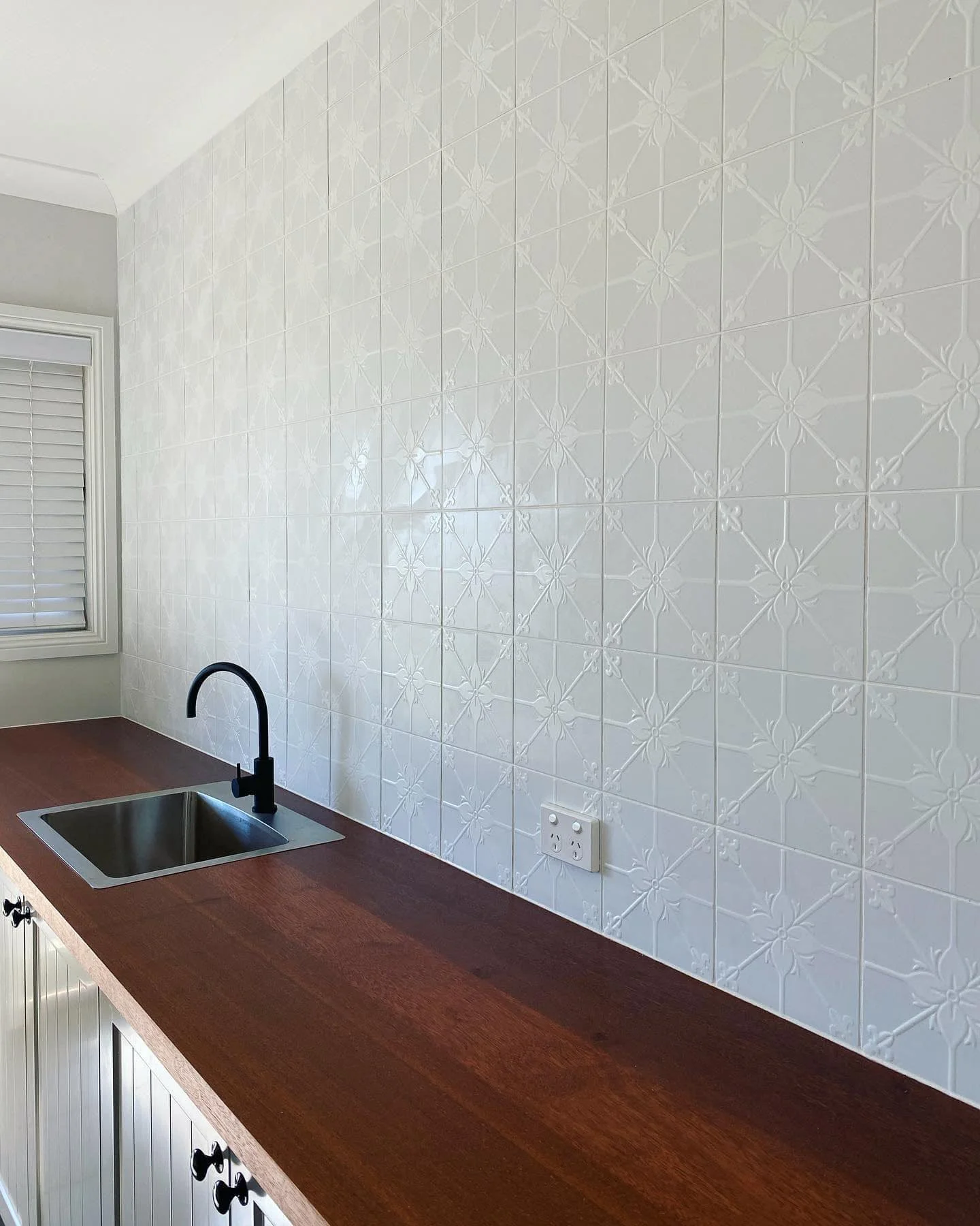 Kitchen countertop with a built-in sink and black faucet, white tiled wall with a decorative pattern, and a window with closed blinds.