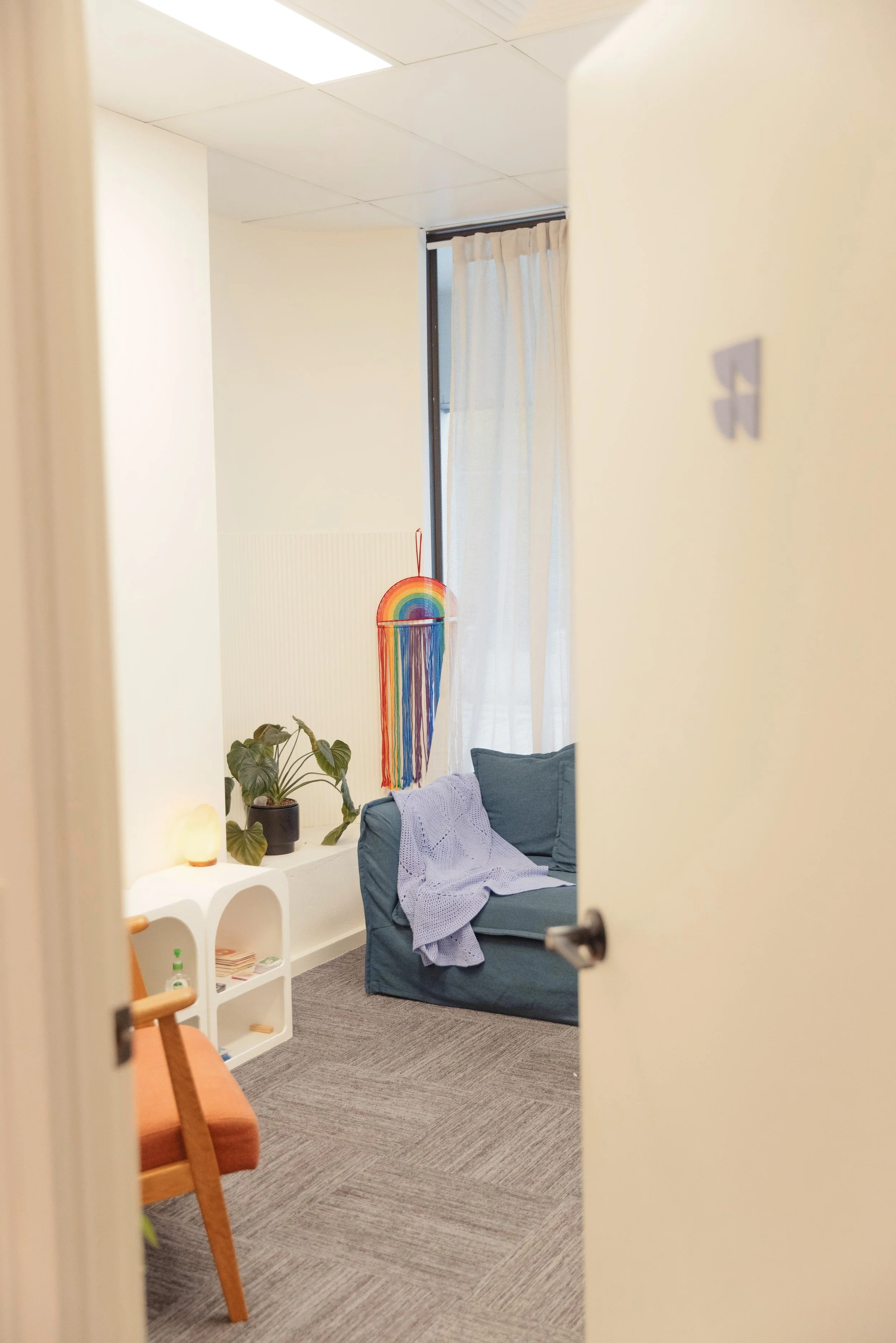 A cozy room seen through a partially open door, with a dark blue couch with a purple blanket draped over it, a potted plant, rainbow-shaped wall hanging, white curtains, and a small white side table.