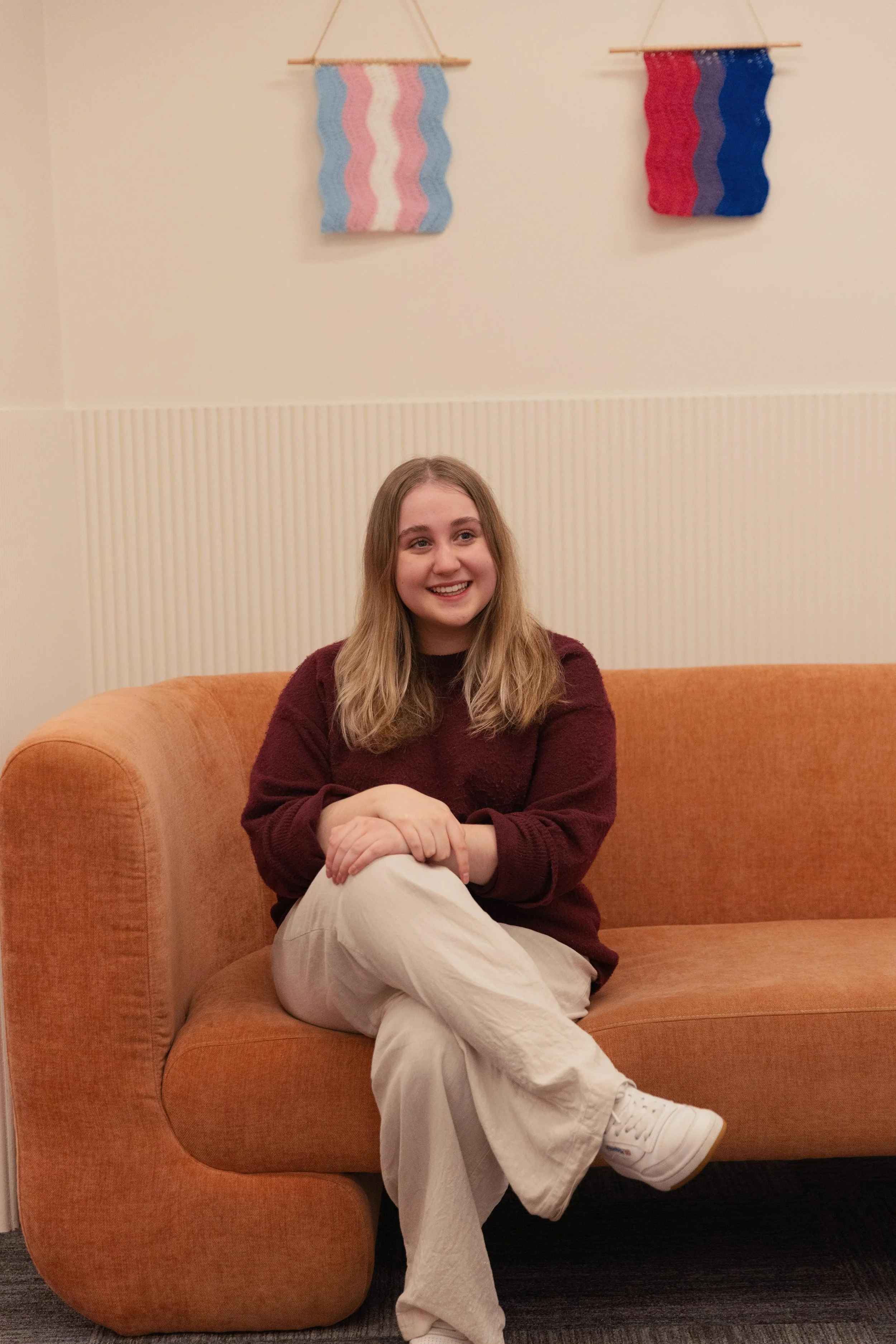 A young woman with shoulder-length blonde hair is sitting on an orange sofa, smiling and looking to her right. She is wearing a maroon sweater, white pants, and white sneakers. Behind her, two colorful woven wall hangings are displayed on the beige wall.