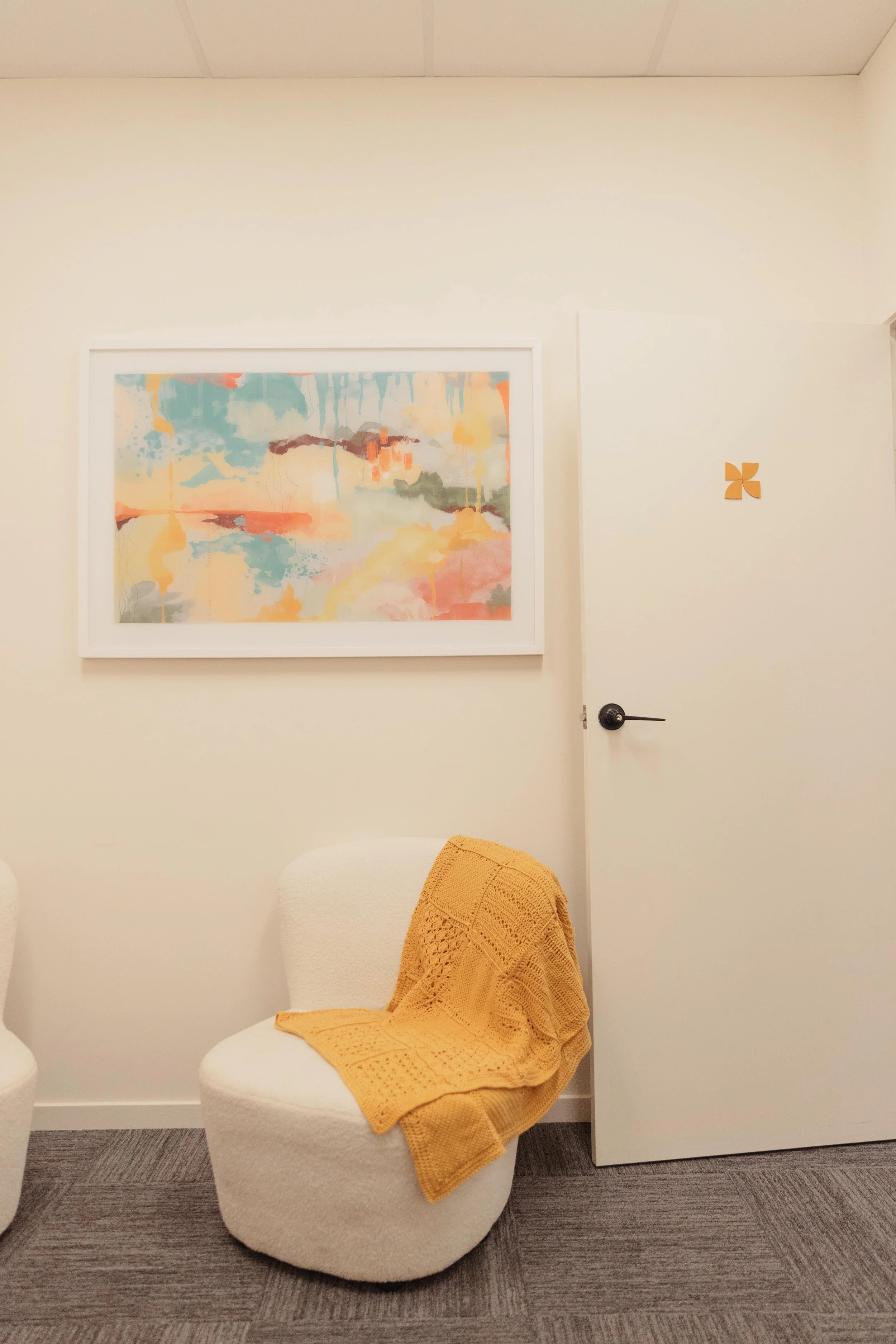 A waiting room corner with a white accent chair, a yellow knitted throw blanket, a colorful abstract artwork on the wall, a white door with a yellow pinwheel design, and a gray carpeted floor.