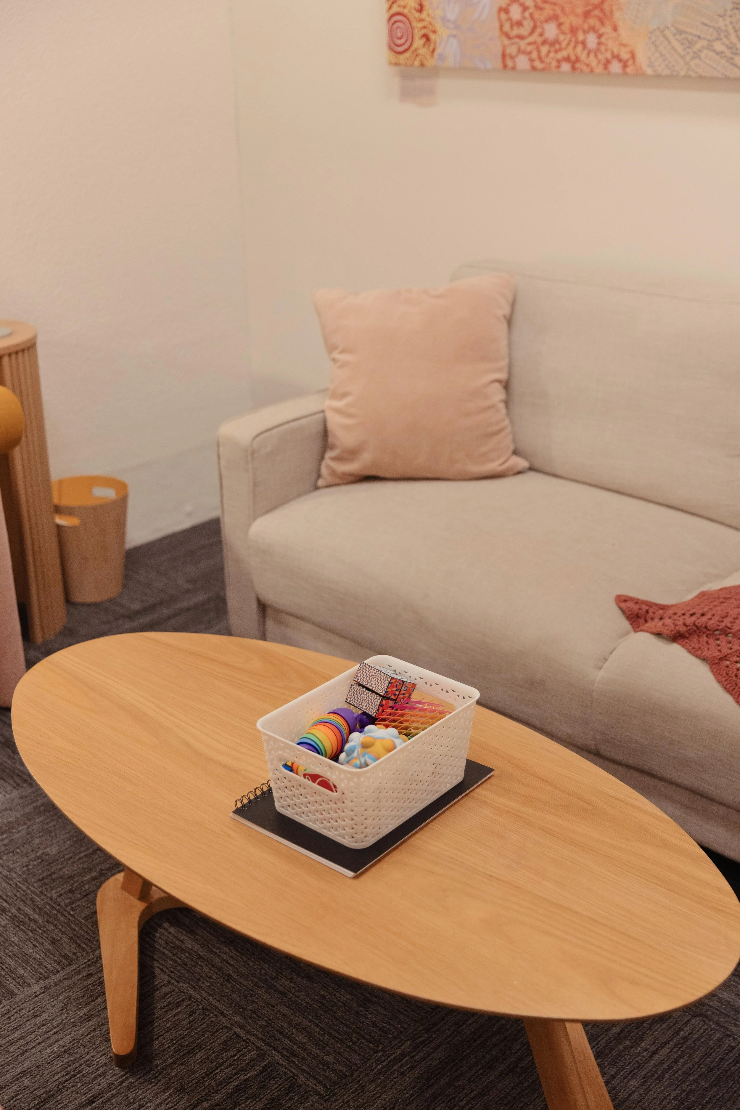 Living room with a beige sofa, pink cushion, and a wooden coffee table with a white basket filled with colorful toys and puzzles.