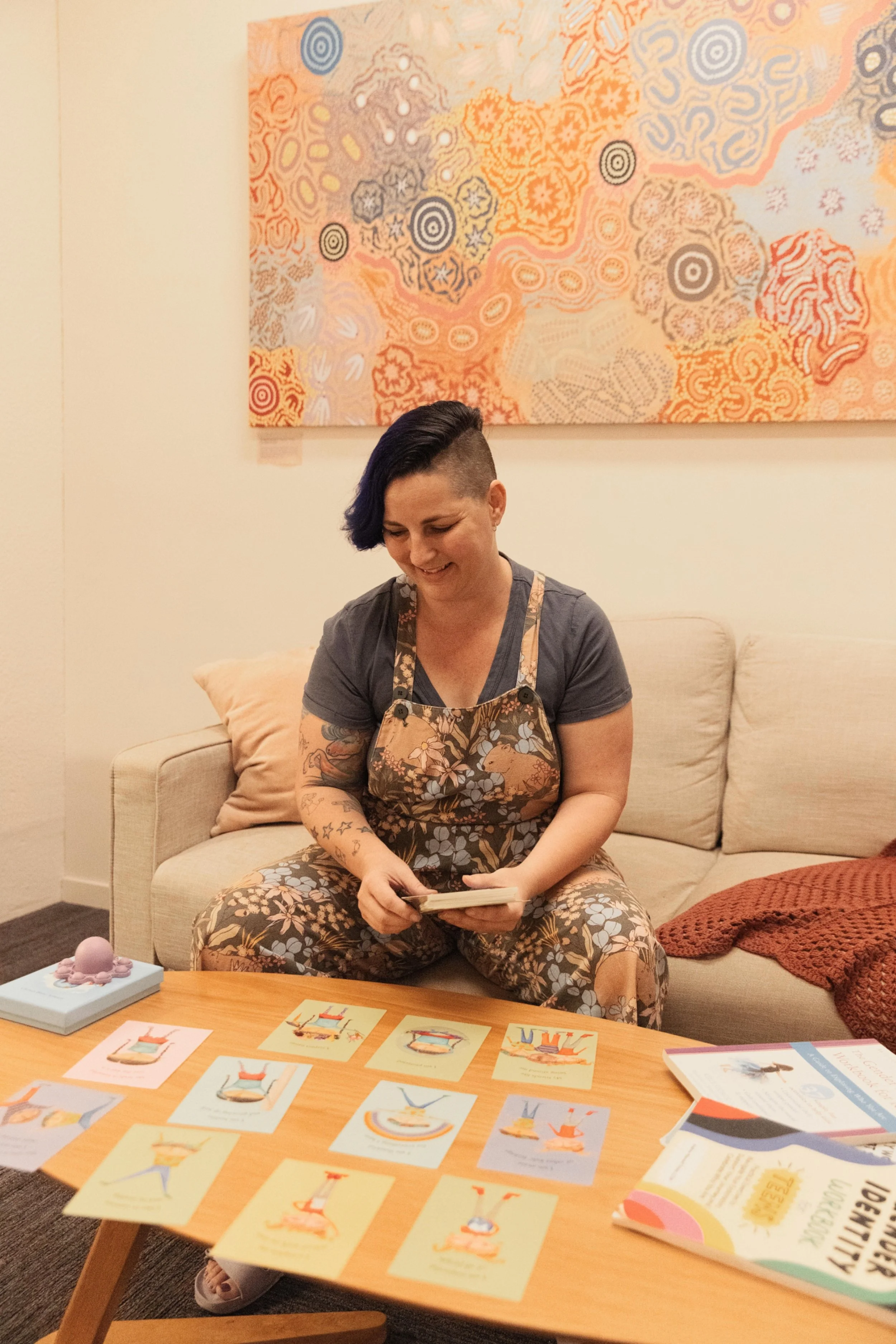 A woman with short dark hair and tattoos on her arms is sitting on a beige sofa, smiling while looking at a stack of cards. She is wearing a vibrant, patterned apron over a grey t-shirt. There are colorful cards with illustrations on a wooden table in front of her, a purple decorative object to the left, and an open book on the right. Behind her, a large abstract painting with orange, peach, and blue patterns hangs on the wall.