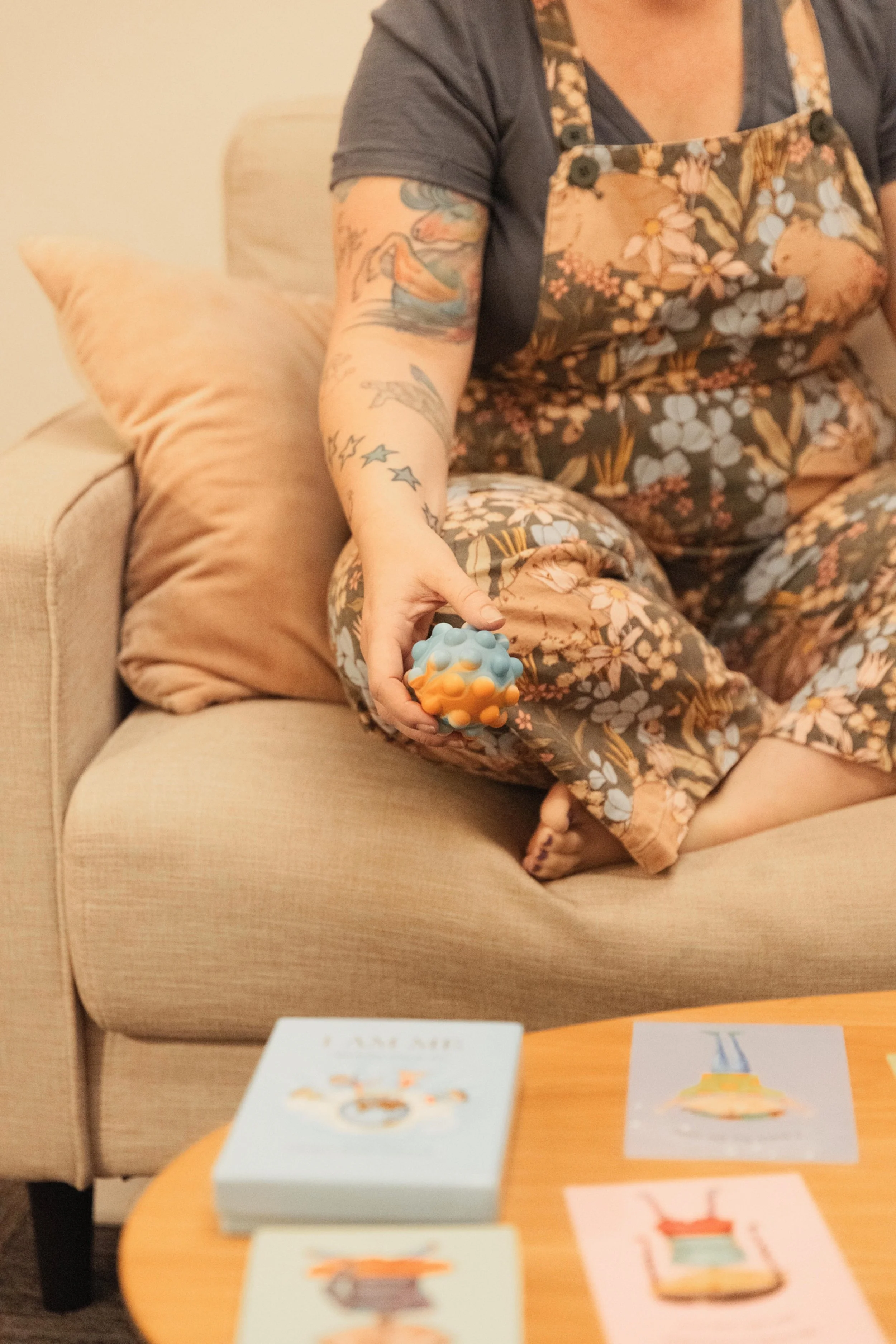 A person sitting on a beige sofa holding a textured therapy ball in their right hand. The person is wearing floral pajamas and has tattoos on their arms. There are pastel-colored cards on a wooden table in front of them.