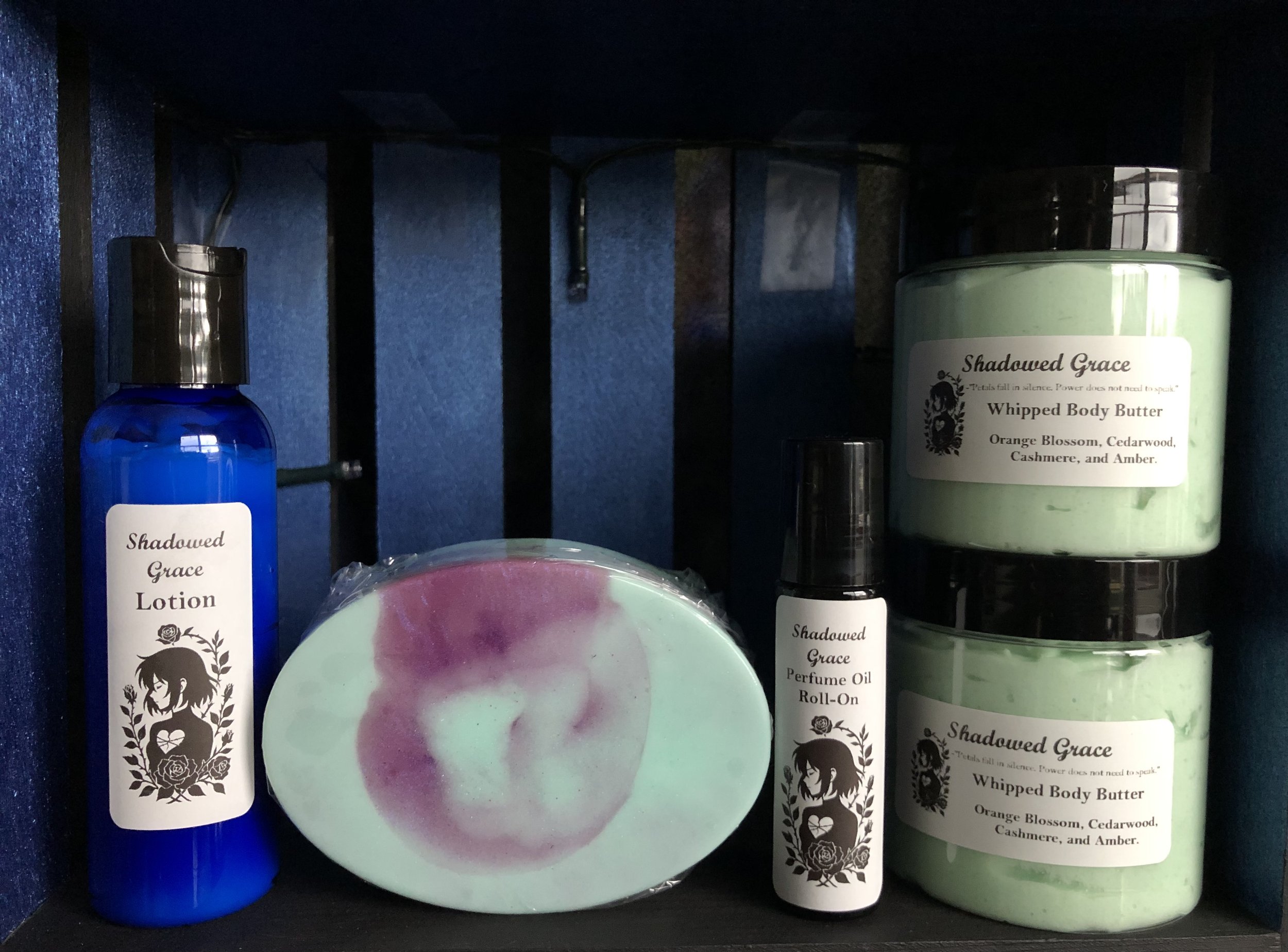 Skincare and body products on a black shelf, including a blue bottle labeled 'Shadowed Grace Lotion,' a round soap with a purple and green swirl pattern, a small perfume oil roll-on labeled 'Shadowed Grace,' and two stacked jars labeled 'Shadowed Grace Whipped Body Butter' with scents of orange blossom, cedarwood, cashmere, and amber.