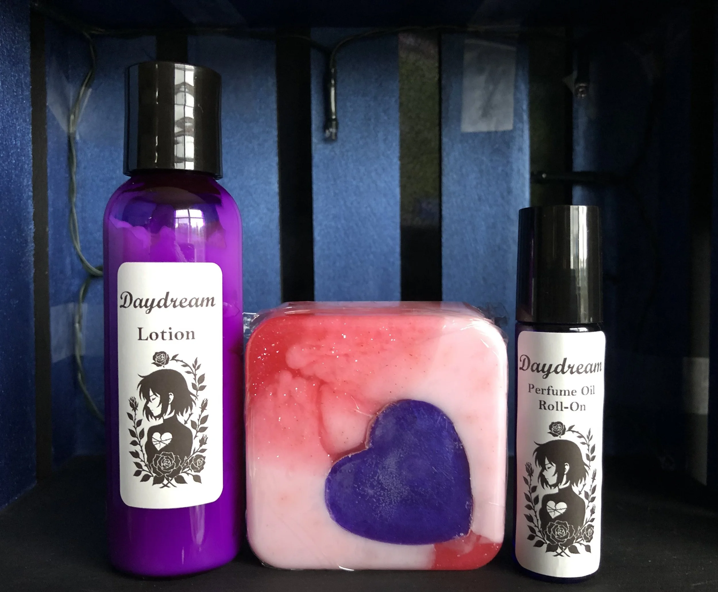 A set of personal care products on a black shelf, including a purple bottle labeled 'Daydream Lotion,' a soap with a purple heart design, and a small spray bottle labeled 'Daydream Perfume Oil Roll-On.'