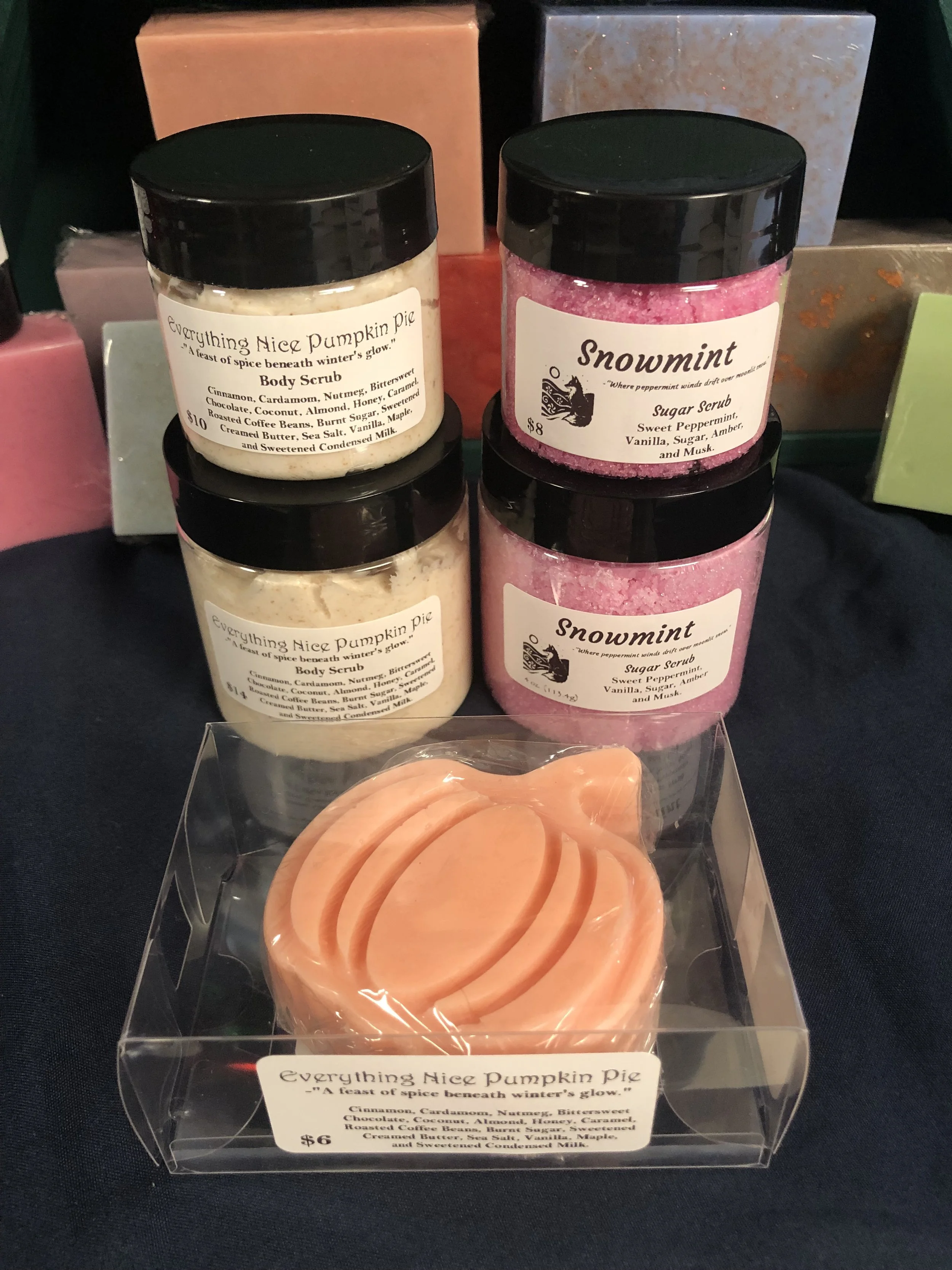 Two jars of body scrub labeled "Everything Nice Pumpkin Pie" and two jars of sugar scrub labeled "Snowmint" behind a clear plastic container of peach-colored soap slices. All items are placed on a dark fabric surface.
