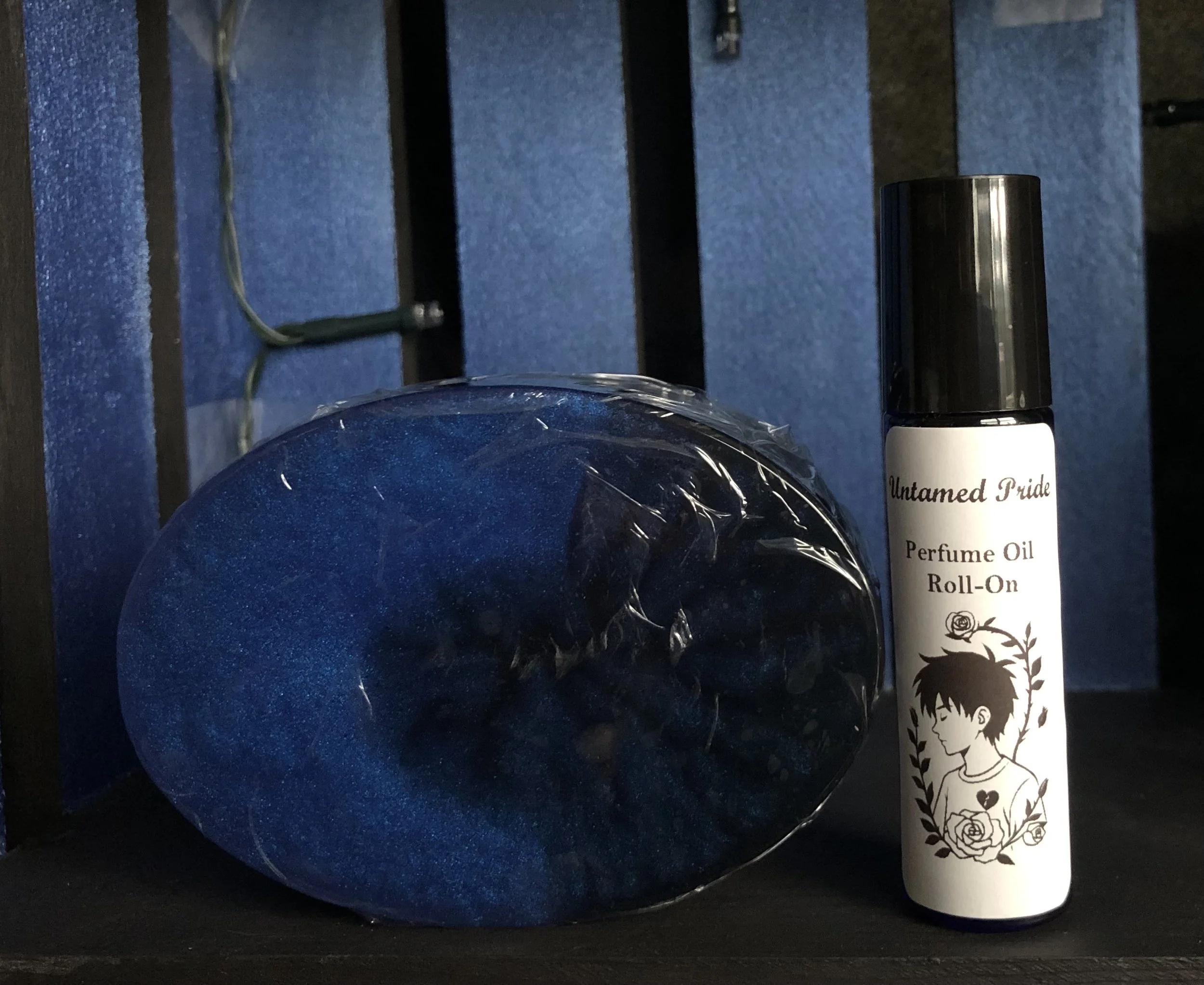 A blue oval soap bar wrapped in plastic and a white perfume oil roll-on bottle with a black cap, bearing a label with an illustration of a person and flowers.