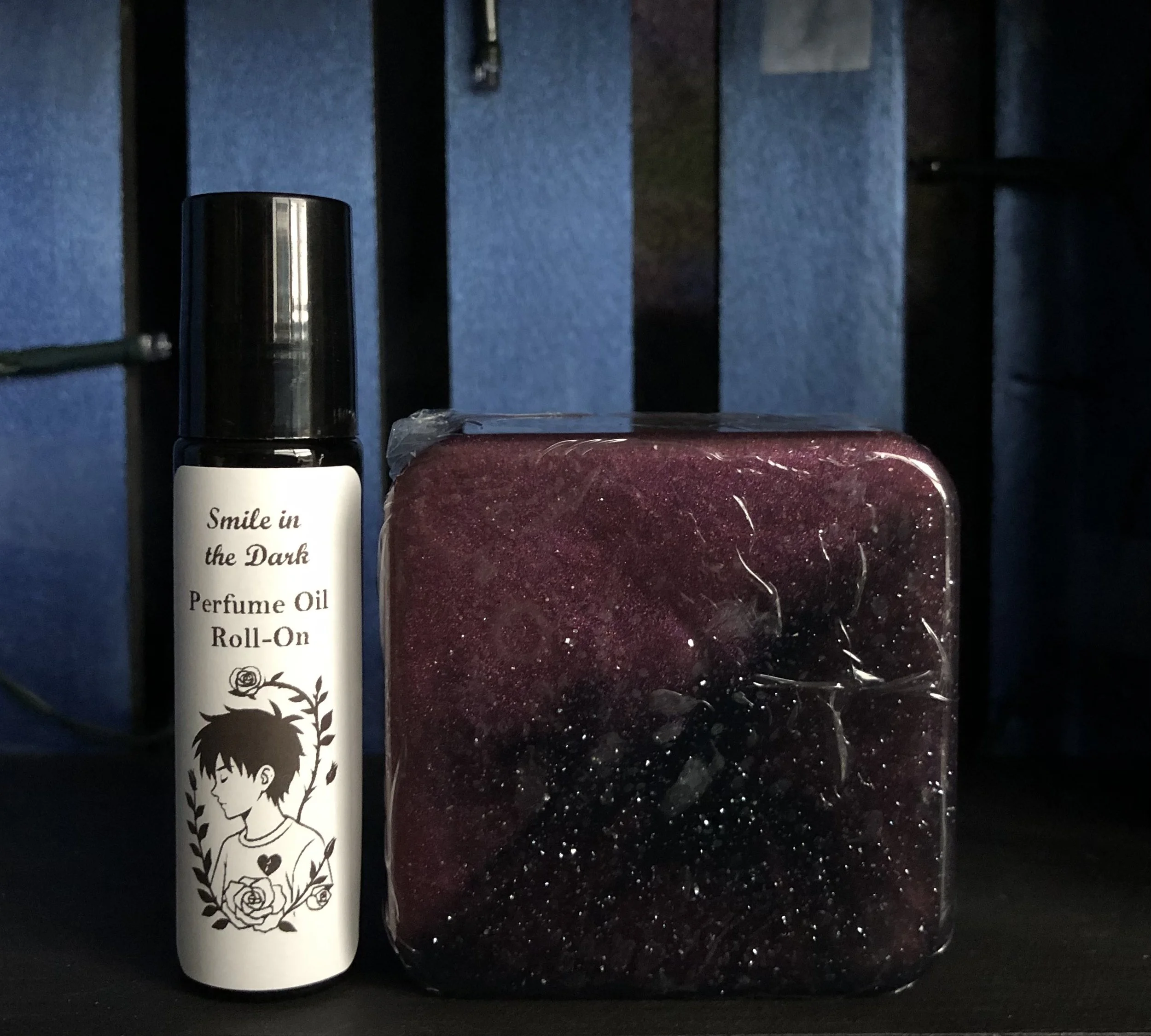 A black spray bottle labeled 'Smile in the Dark Perfume Oil Roll-On' with an illustration of a boy and floral design, placed next to a square container filled with dark purple and black glittery substance.
