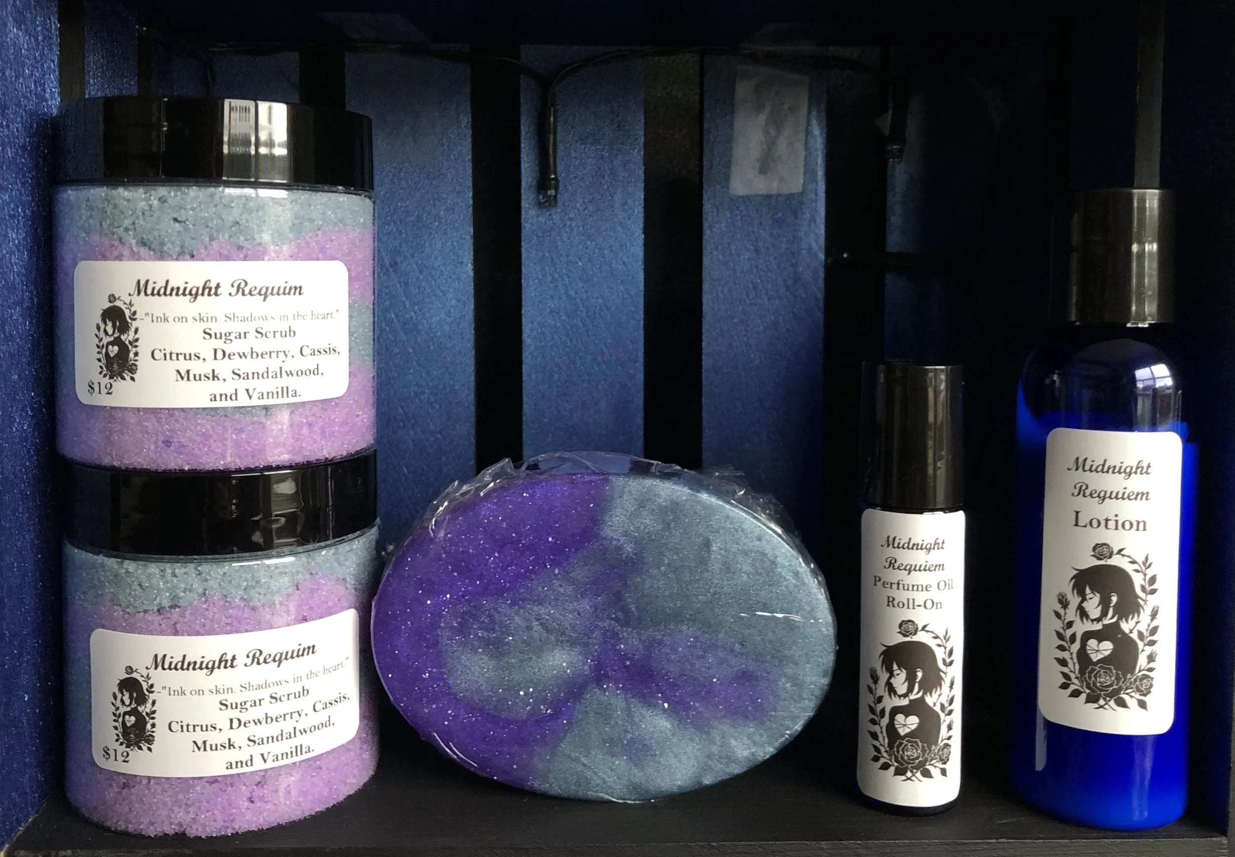 A black shelf holding three purple sugar scrub containers labeled 'Midnight Requiem' with ingredients and a price of $12, a round soap with a swirl of purple and green glitter, a small perfume oil spray labeled 'Midnight Requiem Perfume Oil Roll-On,' and a blue lotion bottle labeled 'Midnight Requiem Lotion' with a black and white label featuring a woman and floral design.