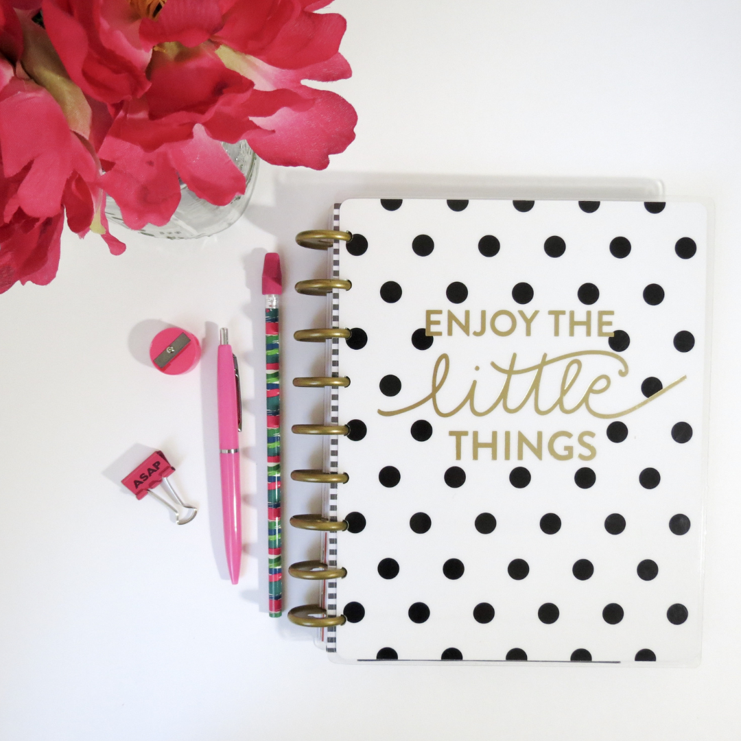 Flat lay of a planner with black polka dots and gold lettering that says 'Enjoy the little things', pink flowers in a vase, a pink pen, a colorful pencil, a small pink sharpener, and a pink 'ASAP' clip on a white surface.