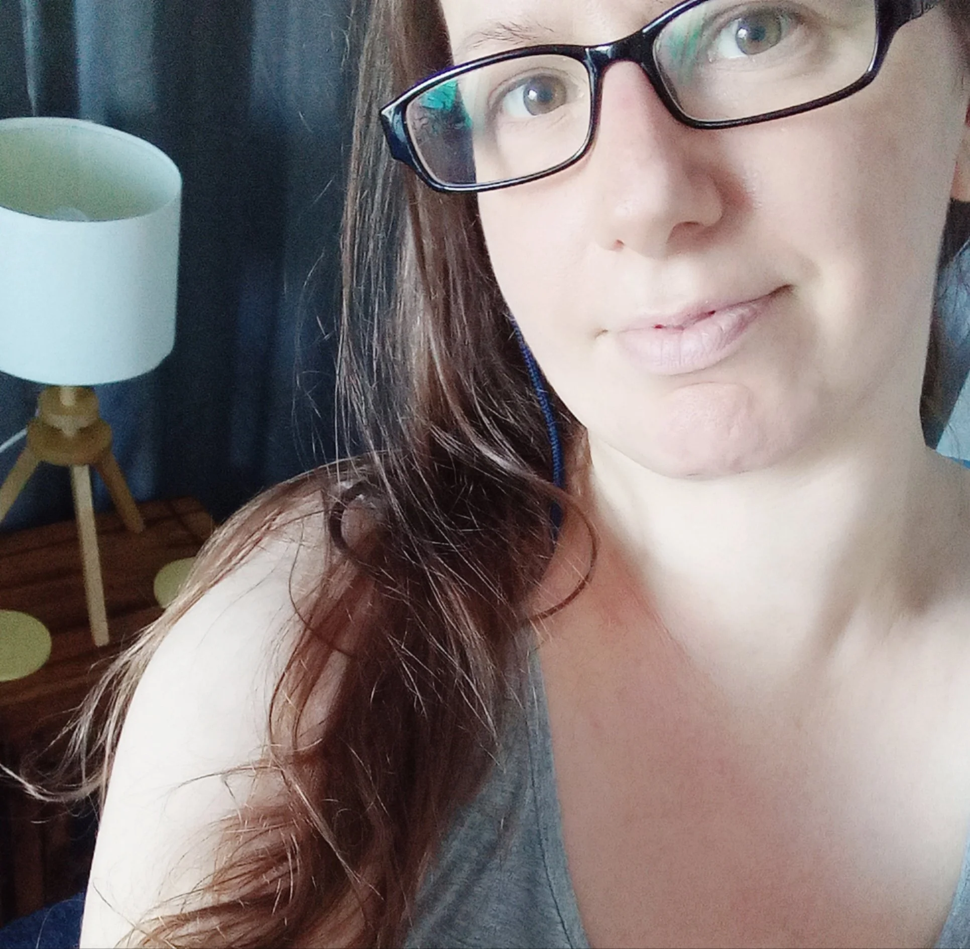 Close-up of a woman with glasses, long brown hair, light skin, and a slight smile, sitting in a room with a white and wooden lamp in the background.