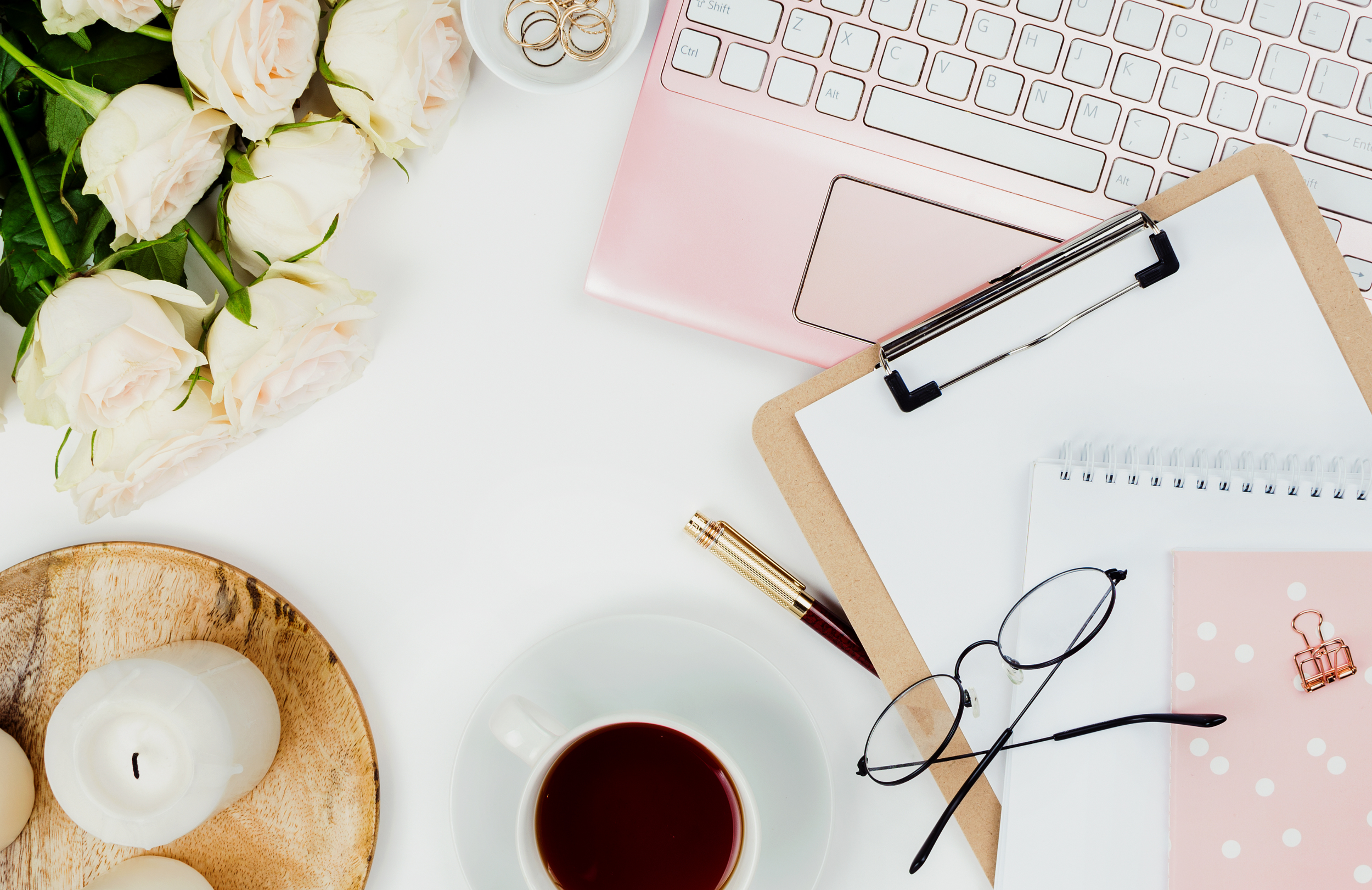 A workspace with a bouquet of white roses, a cup of coffee, a wooden tray with white candles, a pink laptop, a clipboard with papers, a pen, glasses, a pink notebook with polka dots, a paperclip, and a small bowl with rings, all arranged on a white surface.